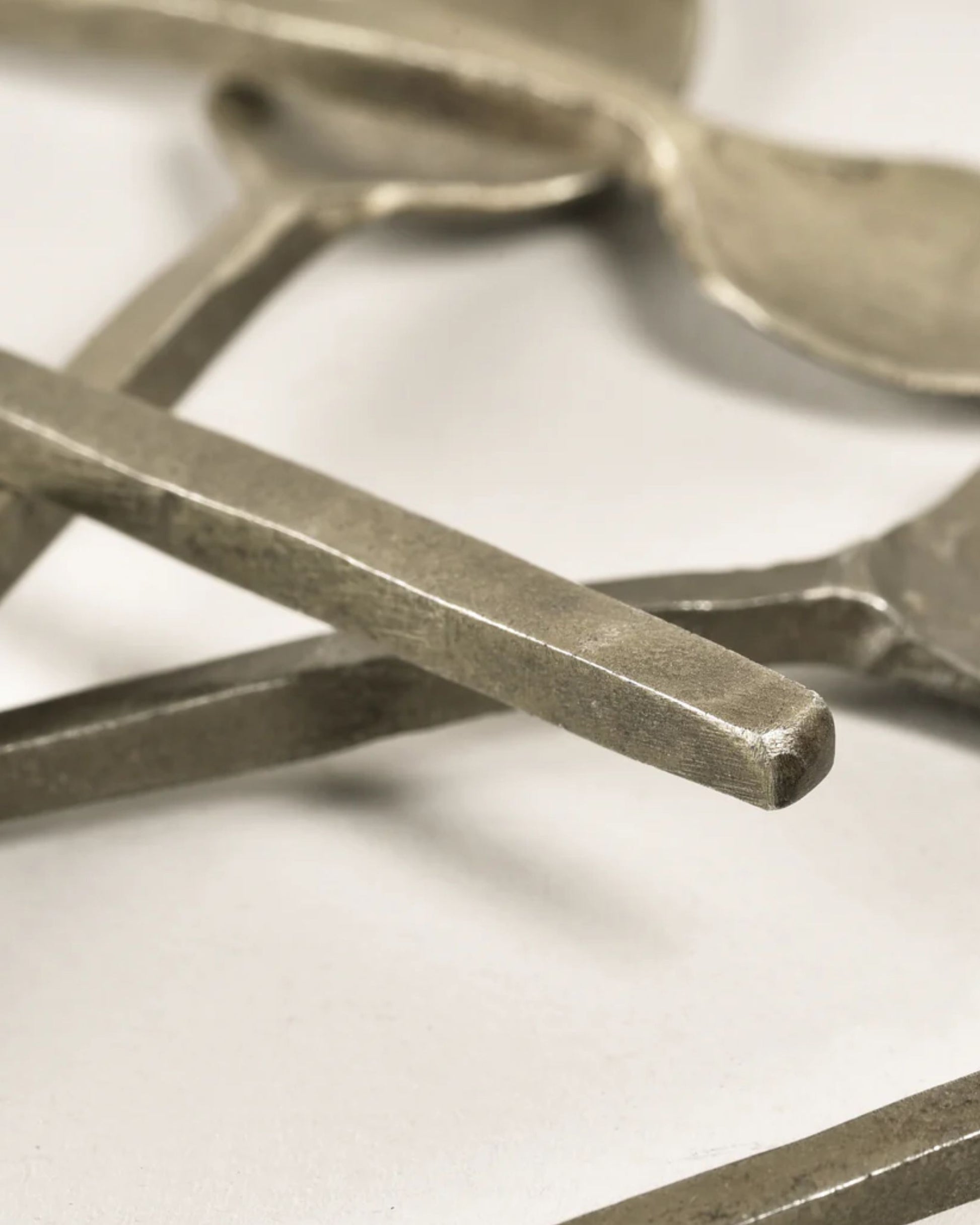 A close-up of the ANTIQUE SALT SPOON set by New Direction, featuring flat, angular handles arrayed casually on a white surface. The brushed metal finish showcases their texture, imbuing each spoon with vintage charm and making them decorative accents in their own right.