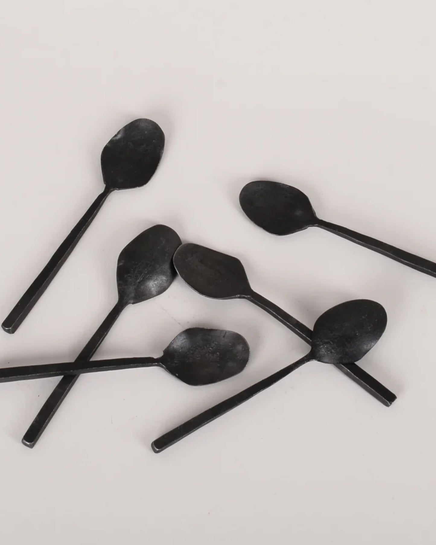 Six ANTIQUE SALT SPOONS from New direction are scattered on a plain white surface, exuding vintage charm. Featuring long handles and slightly rounded ends, each black metal spoon serves as a decorative accent, arranged in a random pattern reminiscent of an antique salt spoon collection.