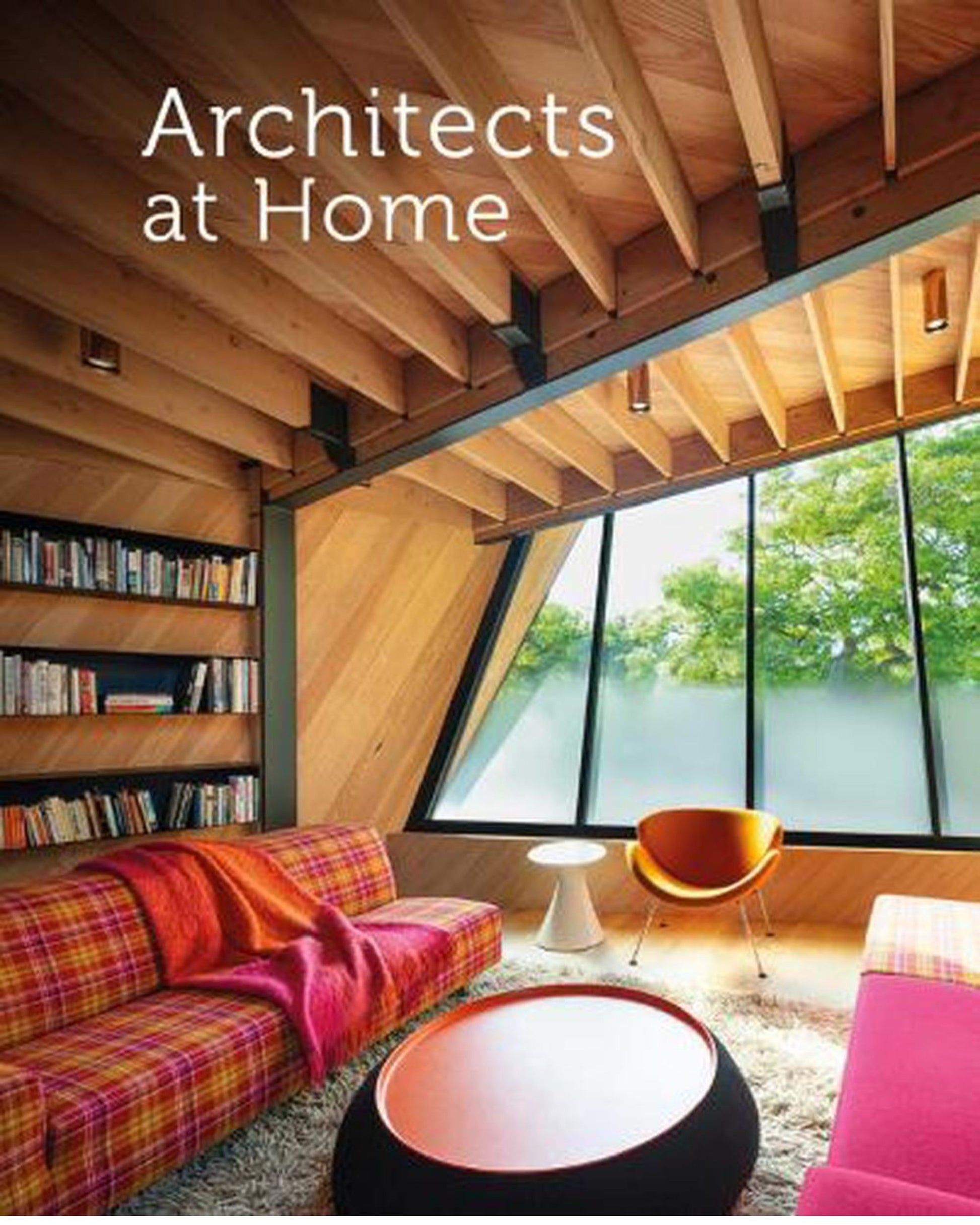 A modern living room with a slanted wooden ceiling and a large window revealing lush greenery embodies the latest design trends. It includes a plaid sofa adorned with a red throw, an orange chair, round tables, and a bookshelf. Text reads: "ARCHITECTS AT HOME" by Nation Wide Books.