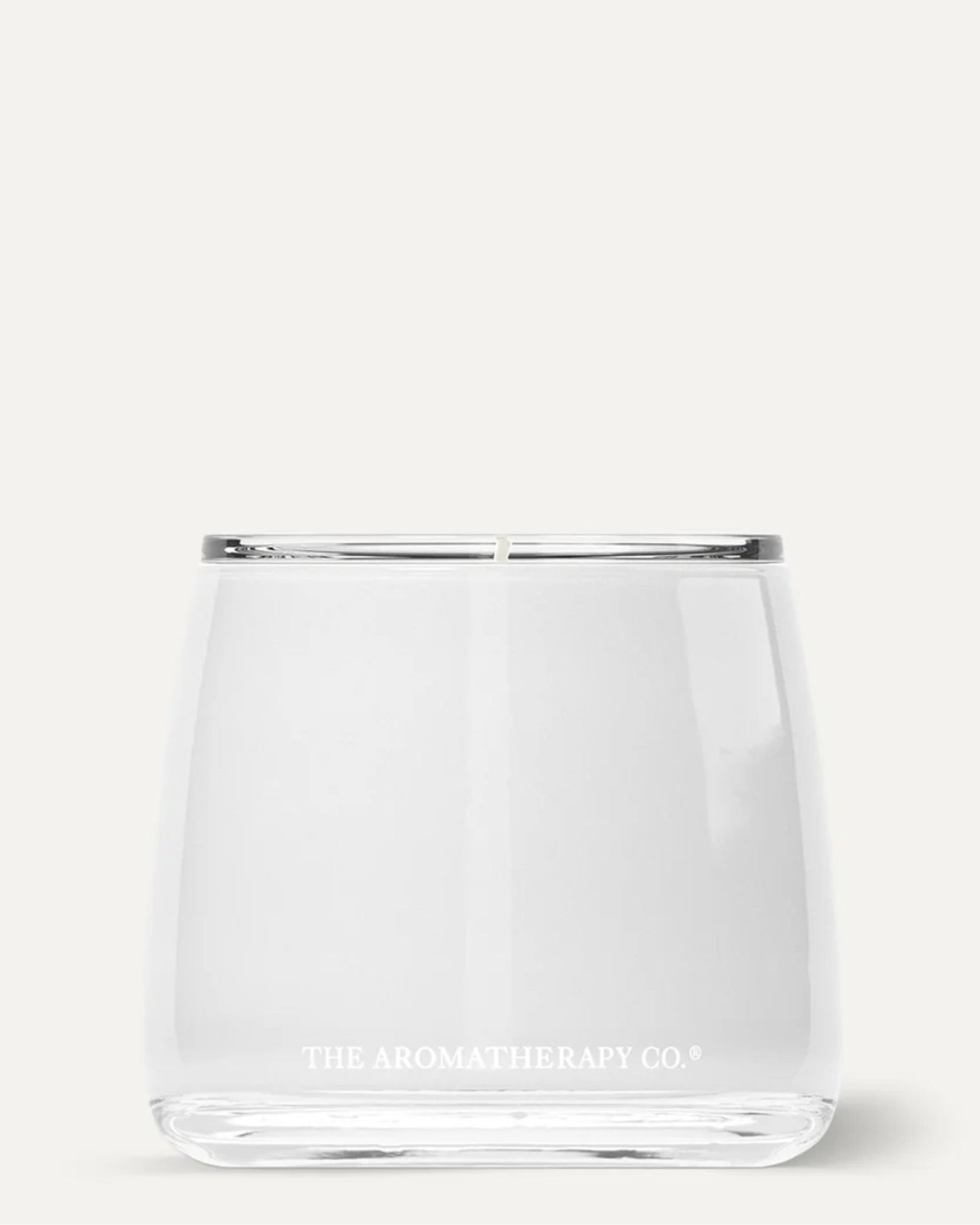 A clear glass candle showcases visible white soy wax, with "The Aromatherapy Co" elegantly inscribed in small white letters near the base. Set against a plain light background, the AROMATHERAPY CANDLE fills any space with calming essential oils, creating a serene ambiance.