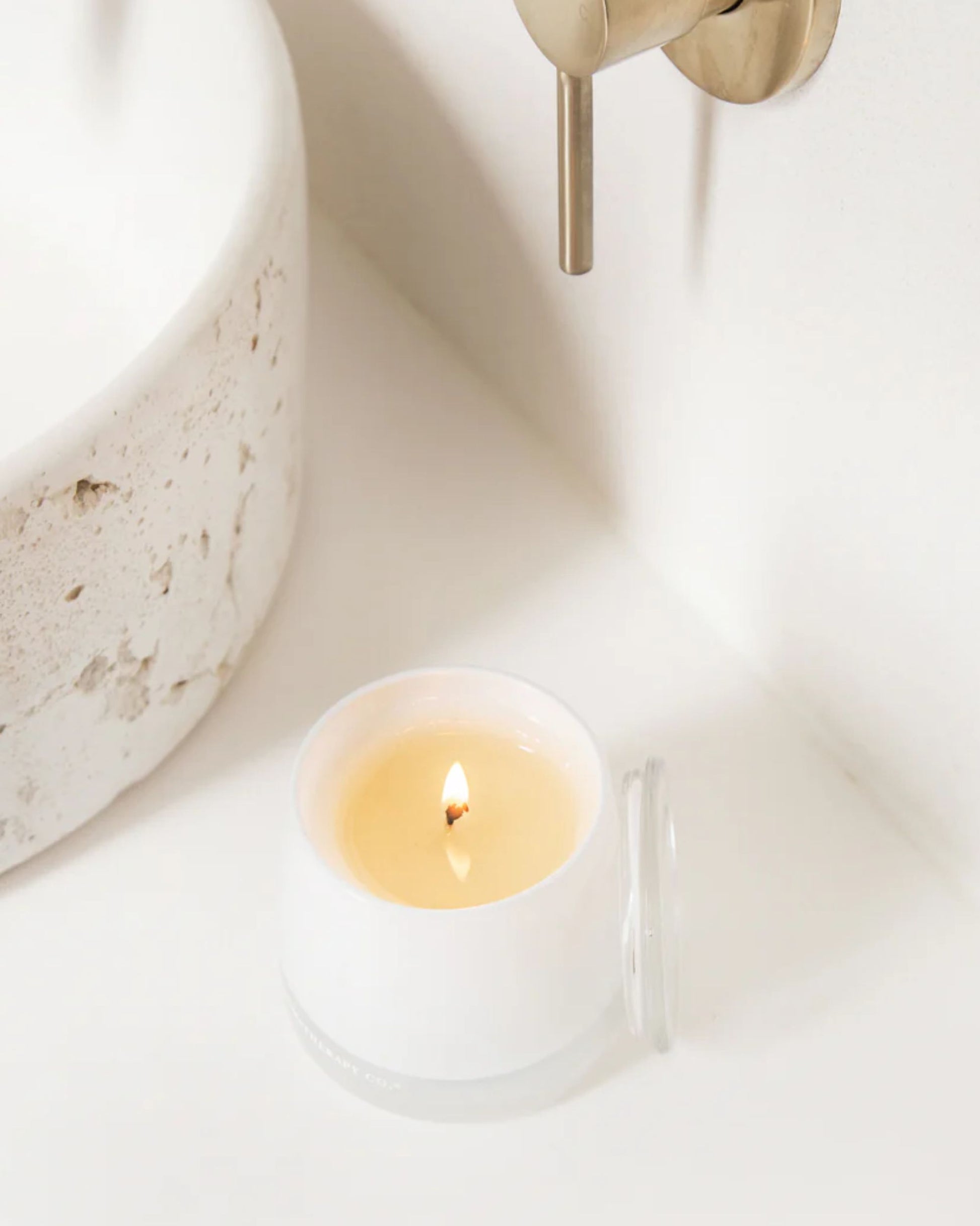 An AROMATHERAPY CANDLE by The Aromatherapy Co sits on a white countertop next to a ceramic sink. Infused with soothing essential oils, its glass jar is open with the lid placed beside it, while a metal faucet is partially visible on the wall.