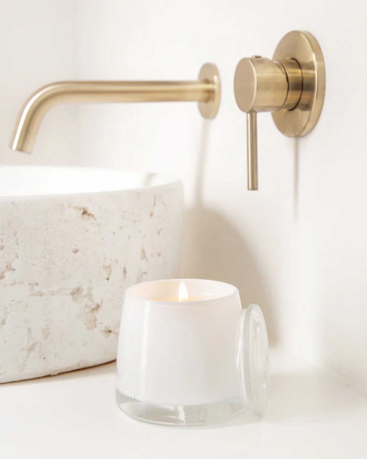 An AROMATHERAPY CANDLE from The Aromatherapy Co emits a gentle glow from its glass holder on the bathroom counter. Nearby, a round granite sink and a modern brass faucet attached to the wall enhance the serene ambiance. Infused with essential oils, this scene evokes a calm and minimalist aesthetic.