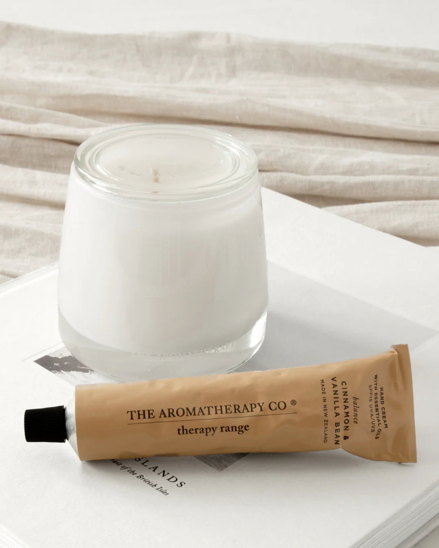 A white candle in a glass holder sits on a white book. In front of the book is a tan tube labeled "AROMATHERAPY HAND CREAM" by The Aromatherapy Co, featuring the therapy range with Cinnamon & Vanilla Bean and enriched with essential oils. A light fabric wraps partially around the book.