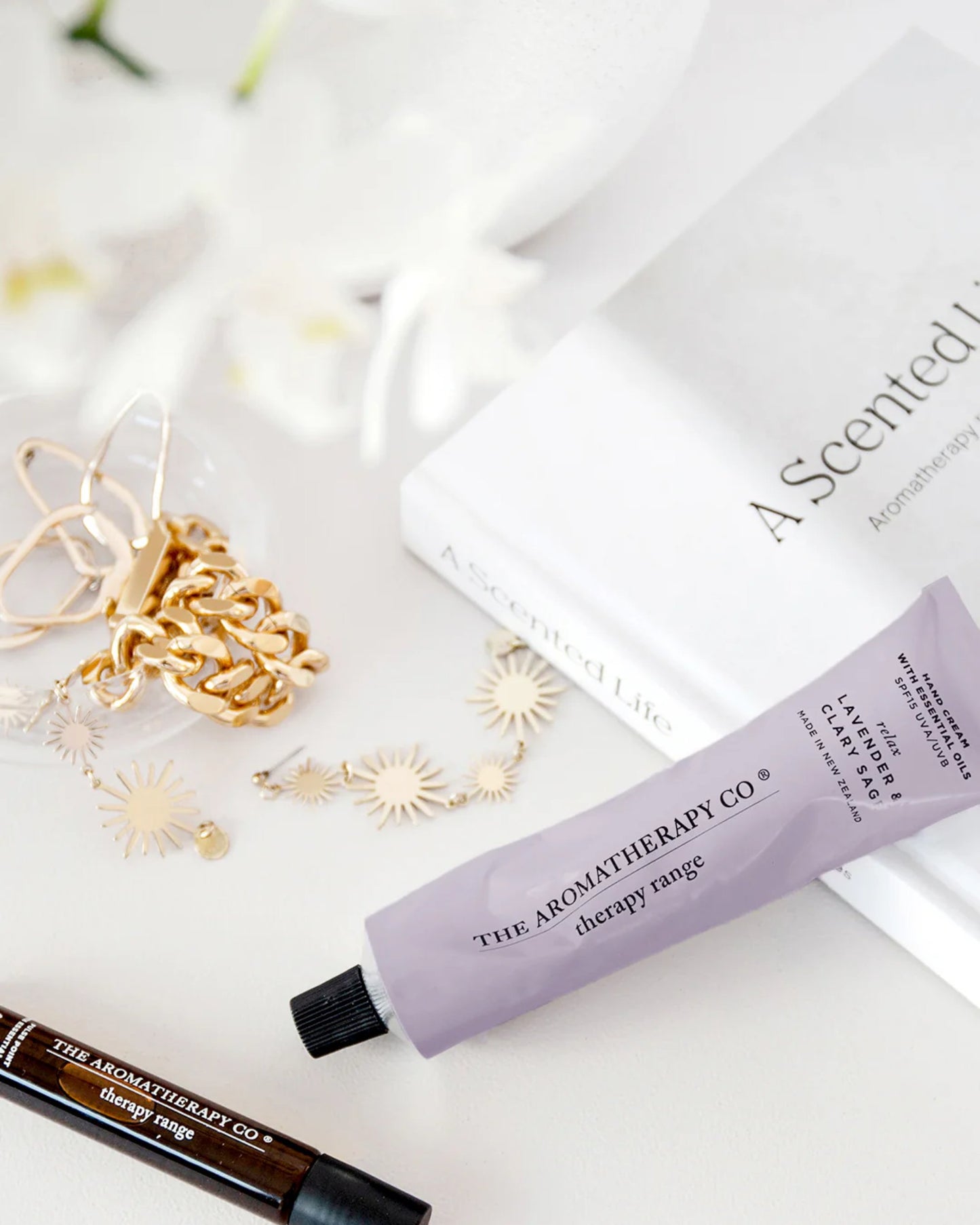 A flat lay features a lavender tube of AROMATHERAPY HAND CREAM by The Aromatherapy Co, placed next to a golden bracelet, earrings, a roll-on fragrance infused with essential oils, and a white book titled A Scented Life. Delicate white flowers subtly adorn the background.