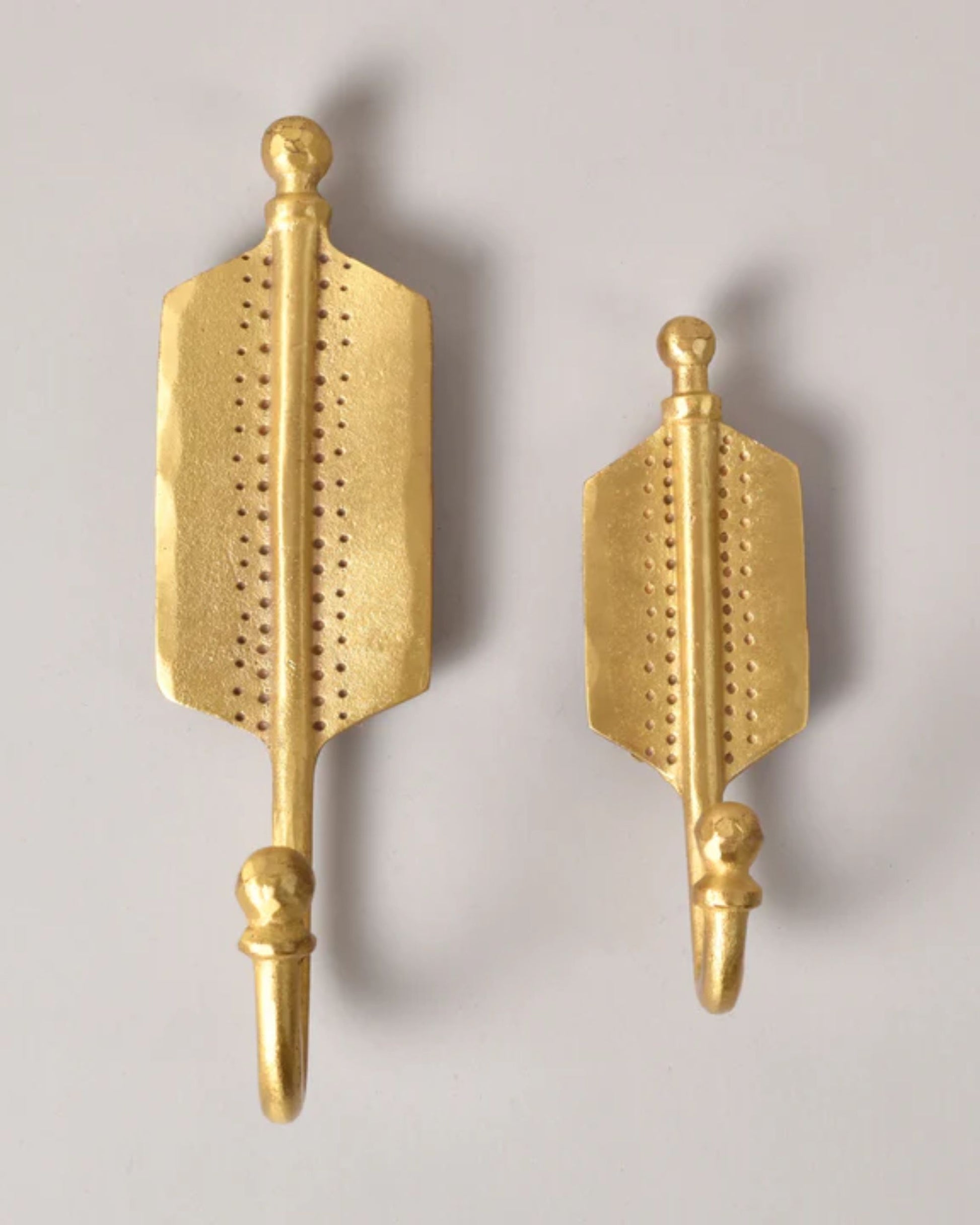 Two ARROW BRASS HOOKS by New Direction adorn the wall, featuring an arrow shape with decorative perforations along their shafts. Available in different sizes, each hook includes a rounded knob on top. These stylish accessories add elegance and practicality to home organization.