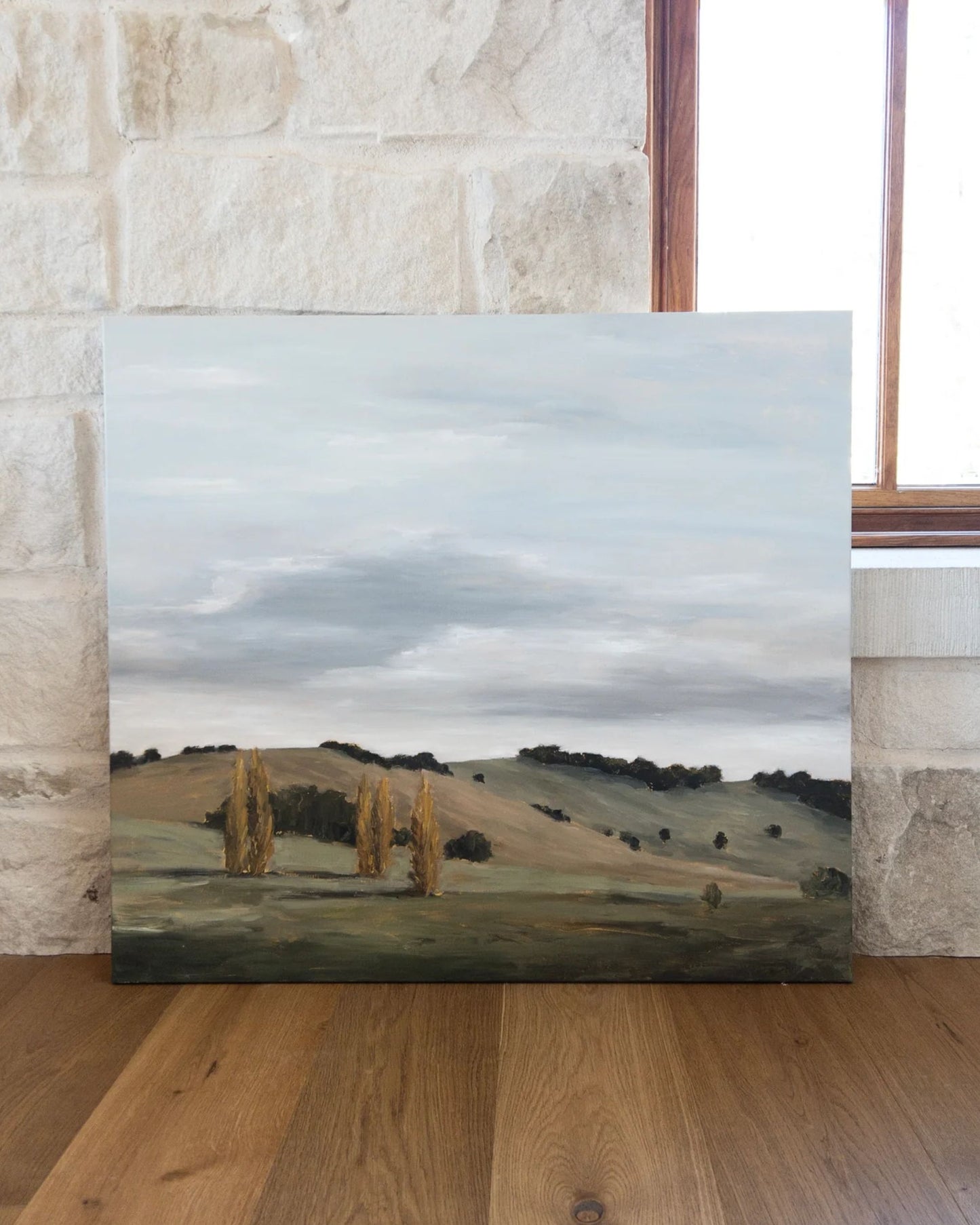 The BURRAWANG DUSK artwork by intuitive artist Jeannie Dolan adorns a wooden floor, displaying rolling hills beneath a cloudy sky. The distant trees and muted earth tones create a serene, pastoral atmosphere. This piece from Art by Jeannie leans against a stone wall, while a window on the right offers glimpses of the natural world.