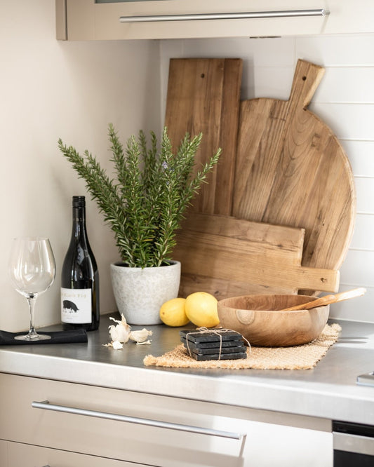 A kitchen counter adorned with a wine bottle, glass, garlic, lemons, and a potted rosemary plant. The Hawthorne Artisan Serving Board (45cm x 25cm) accompanies a wooden bowl and cutting boards, while a wire cheese slicer rests elegantly on a woven mat in the foreground.