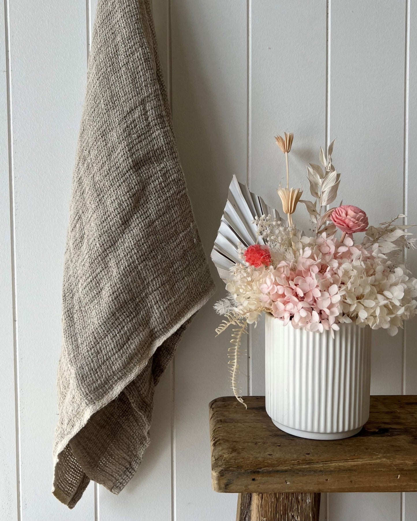 An AUDREY LINEN HAND TOWEL - BONE from Rustic Linen hangs on a white paneled wall. Next to it, a wooden stool holds a white vase filled with dried flowers, such as pale pink and white hydrangeas and palm leaves, creating a rustic, minimalist decor.