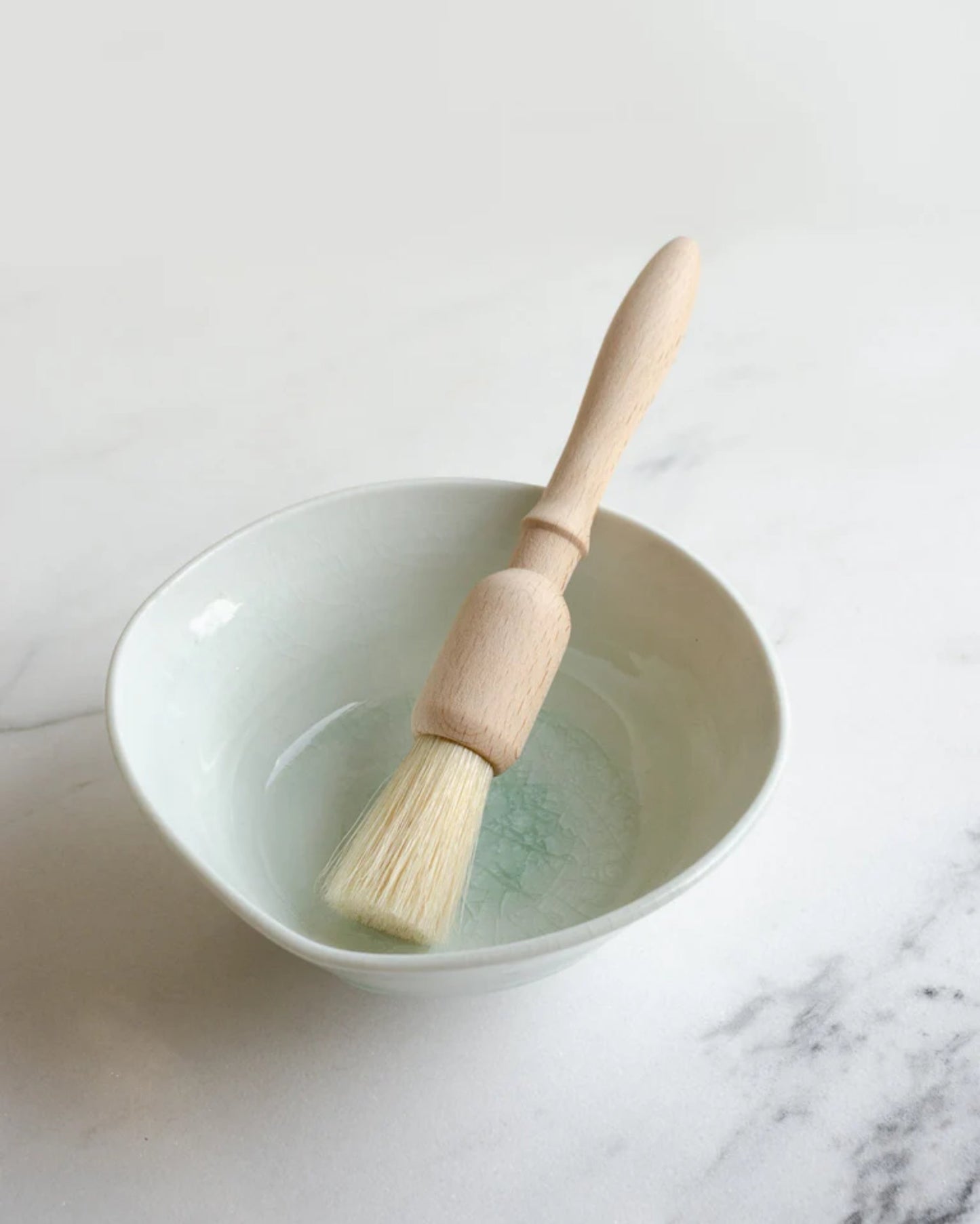 A Redecker BAKING BRUSH - ROUND, crafted from oiled beech wood with natural bristles, rests on the edge of a light gray ceramic bowl filled with water on a white marble surface. The background is softly blurred.