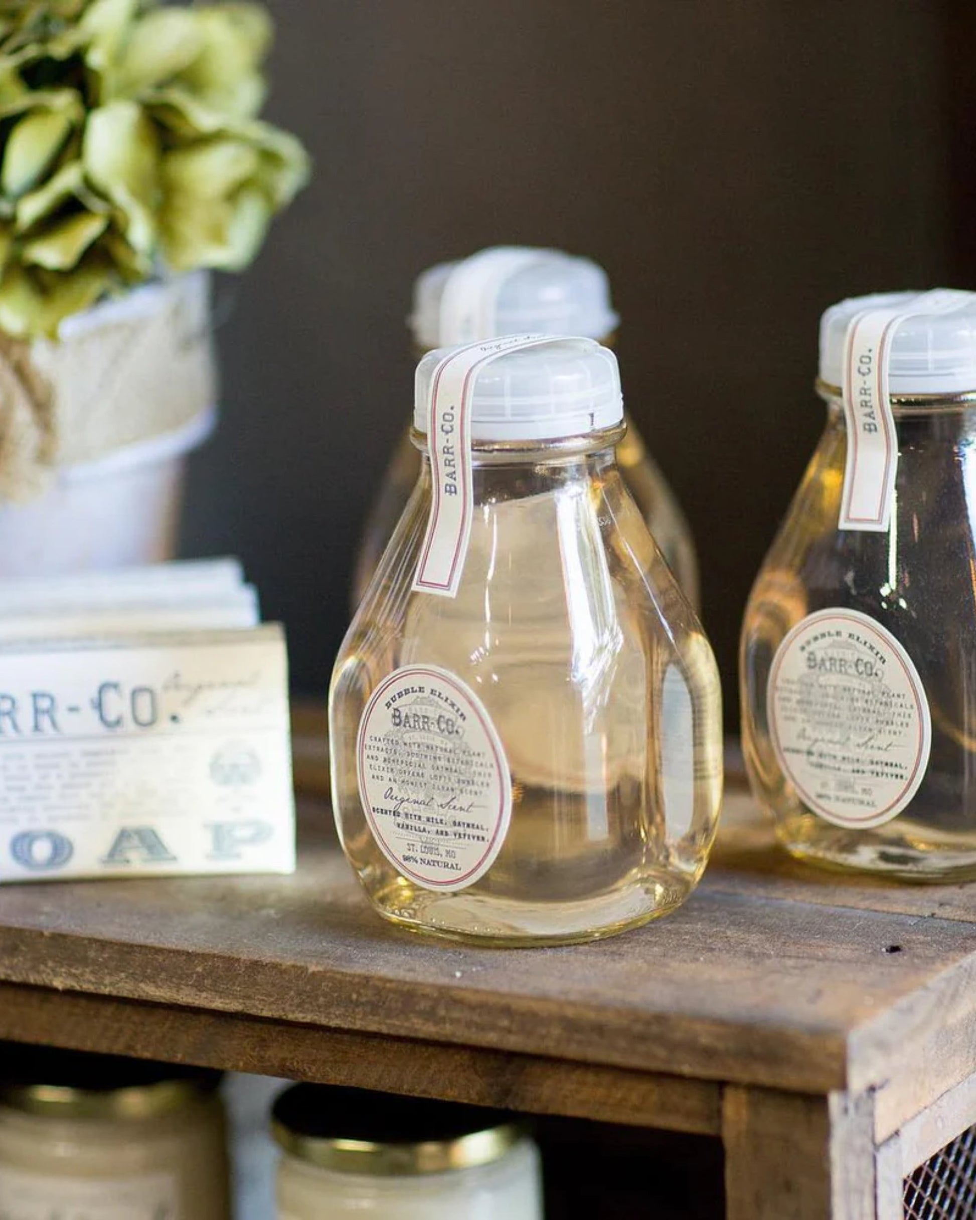 Three BARR CO BUBBLE BATH ELIXIR bottles by Barr Co, made of clear glass and adorned with labels, sit on a wooden shelf, exuding an apothecary-inspired charm. Each bottle holds a yellow liquid, presumably the luxurious bubble bath elixir soap. Also nearby is a potted plant with green leaves and a small bar of soap, enriching the serene tableau.