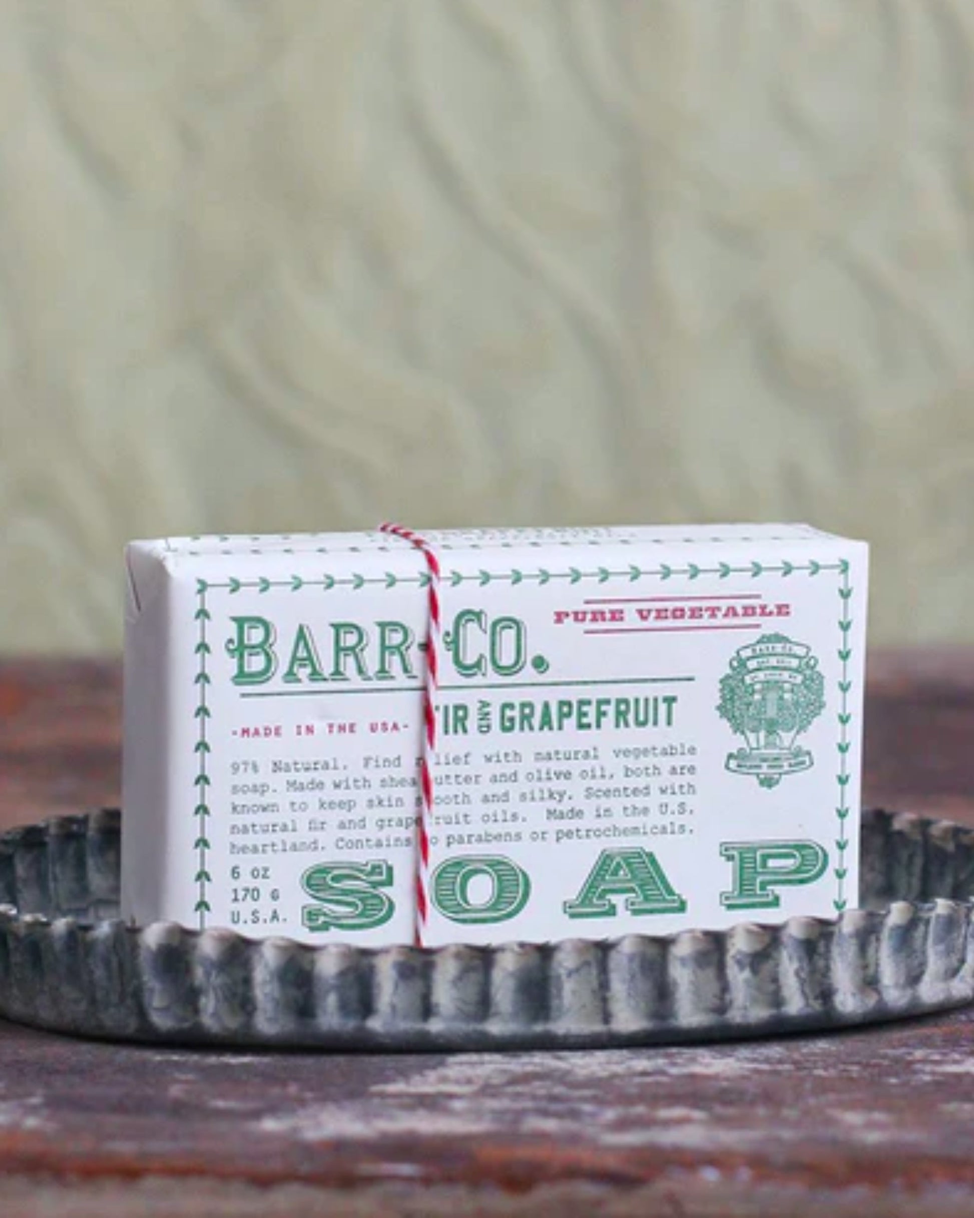 A bar of BARR CO WRAPPED SOAP by Barr Co, featuring a signature fir and grapefruit scent, comes encased in vintage-inspired packaging. It softly exfoliates skin while resting on a rustic, fluted metal tray against a textured backdrop.