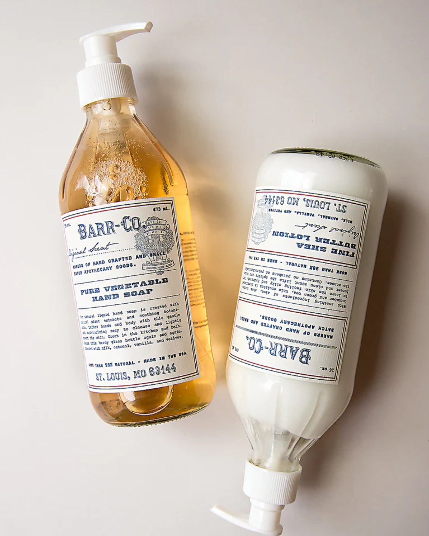 Two bottles of BARR CO LIQUID HAND SOAP are positioned on a surface. One bottle stands upright, filled with amber soap known for its gentle moisturizing properties, while the other rests horizontally, containing a white variant. Their vintage-style labels emphasize the use of natural plant extracts and their origin in St. Louis, MO.