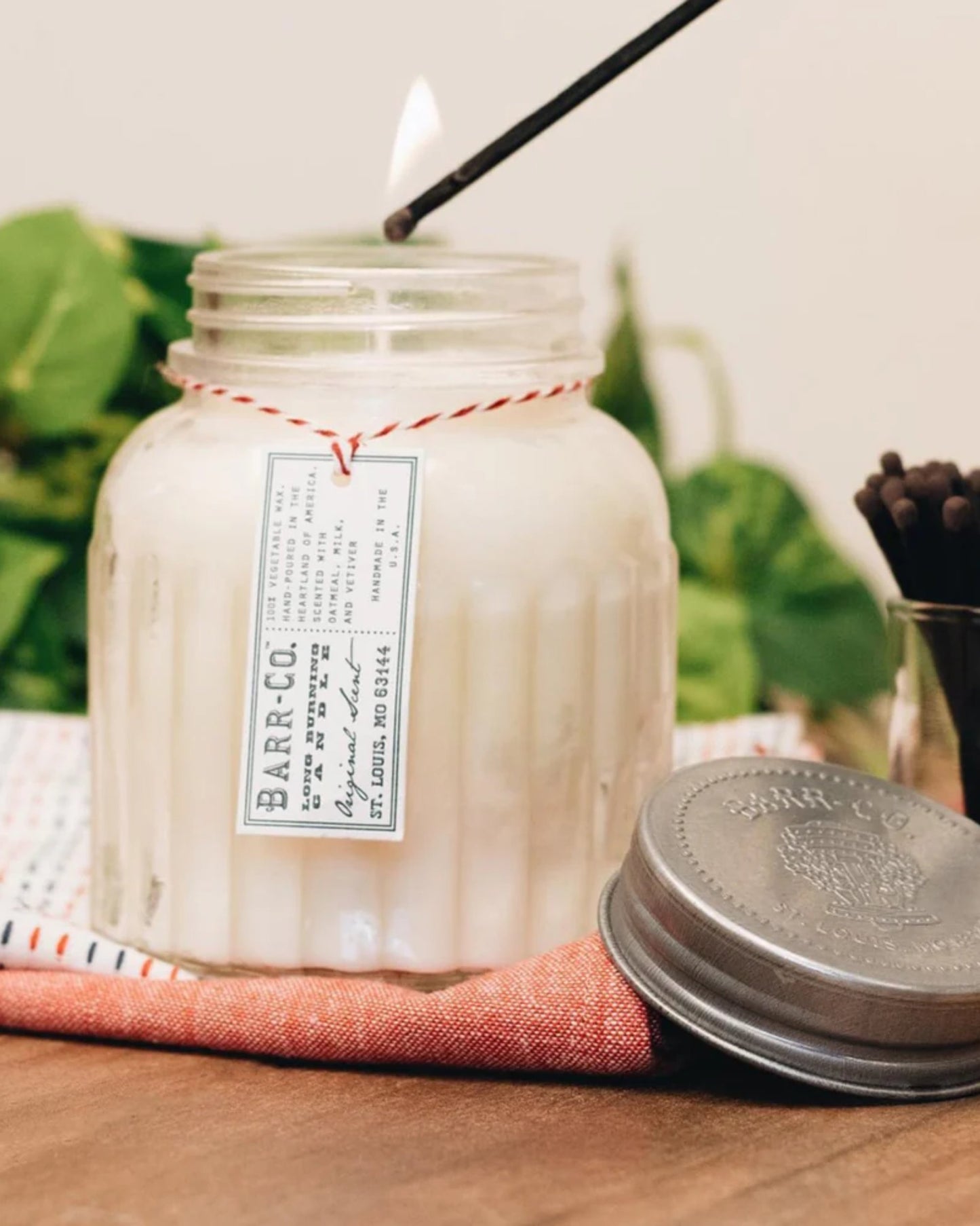 A lit BARR CO ORIGINAL 120HR APOTHECARY CANDLE by Barr Co in a glass jar with a metal lid emits a warm glow on a red cloth as it is lit with a match. The clean-burning soy wax creates an inviting atmosphere, surrounded by green leaves that enhance the cozy ambiance.