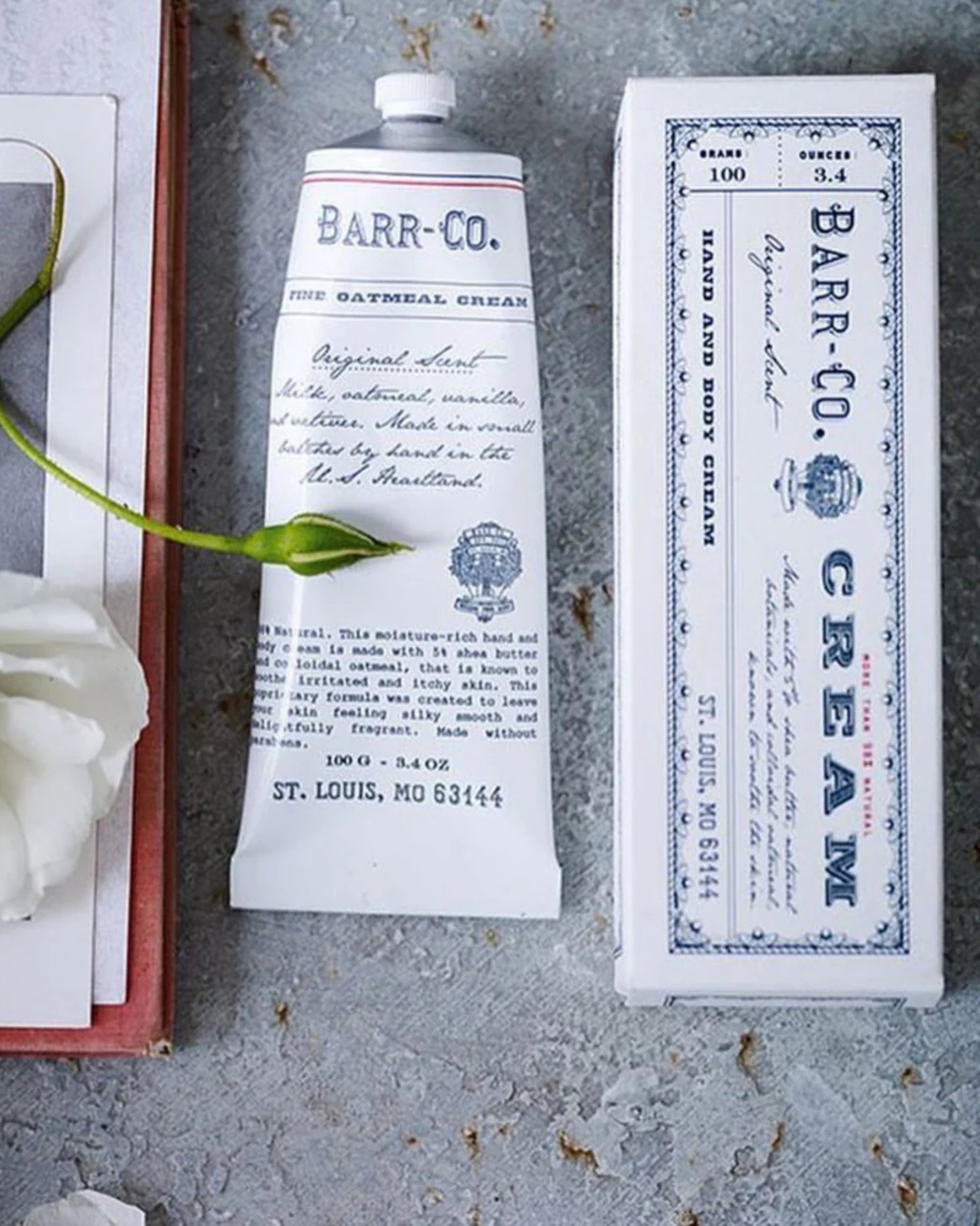 A tube and box of BARR CO ORIGINAL HAND CREAM from Barr Co rest on a textured surface, infused with soothing shea butter. The oatmeal-scented cream showcases a vintage-inspired label, accompanied by a white flower and the edge of a book on the left.