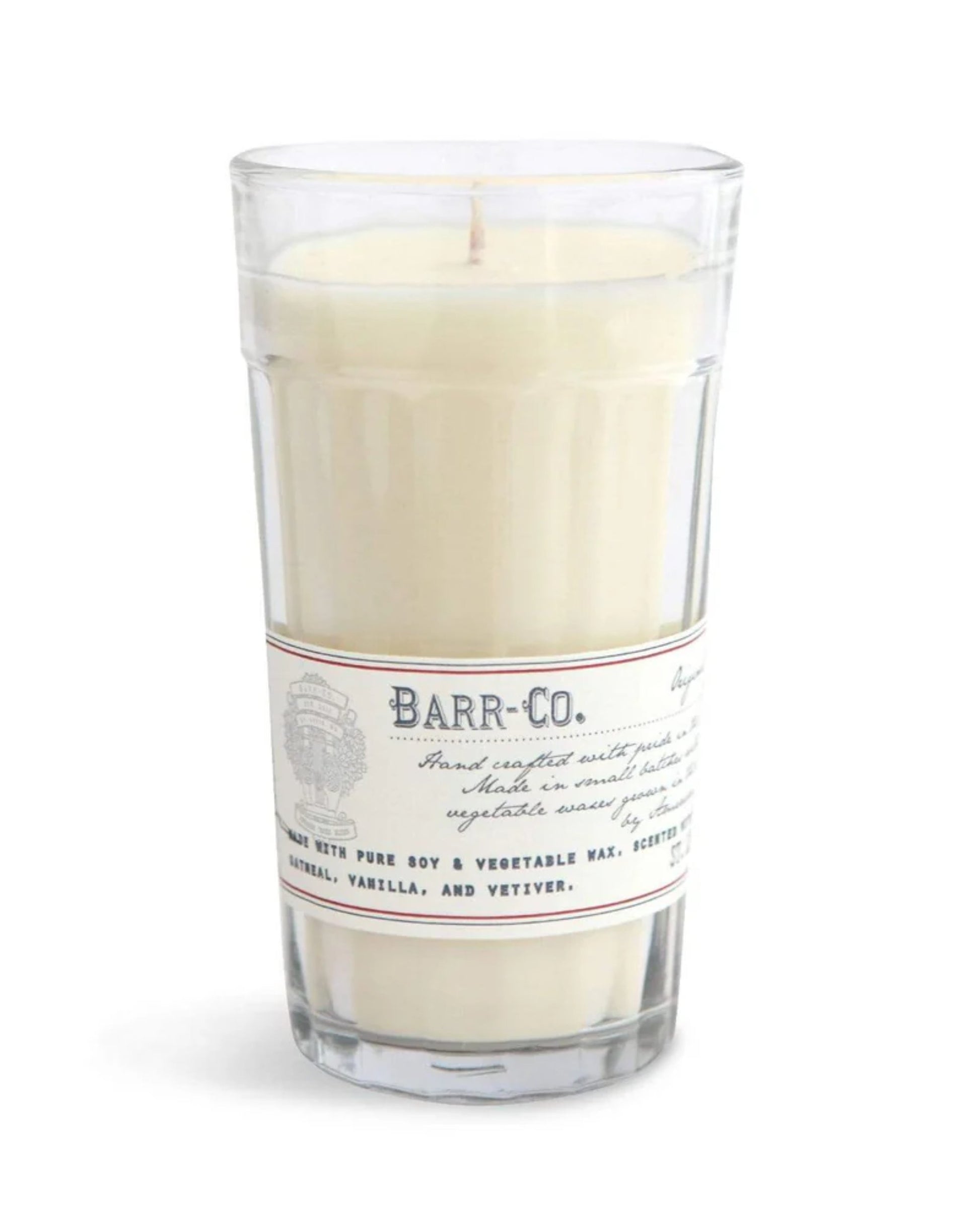 A tall clear glass houses a white candle labeled Barr Co, offering the soothing scents of oatmeal, vanilla, and vetiver. This BARR CO MILK GLASS CANDLE is made from pure soy and vegetable waxes, providing a gentle ambiance suitable for any setting.