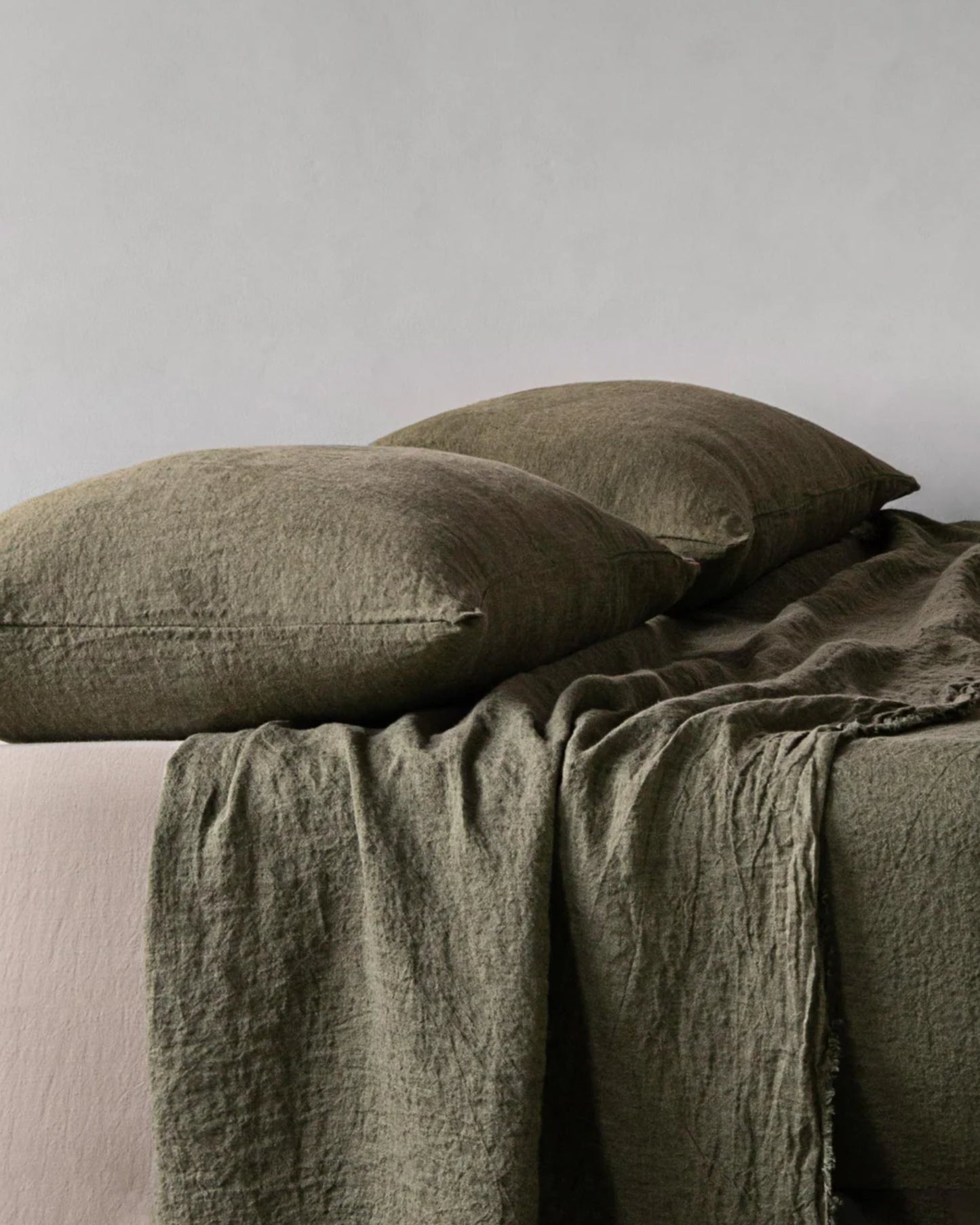 A bed adorned with olive green BASIX STANDARD PILLOW CASES by Hale Mercantile Co and a matching blanket draped over an off-white sheet, set against a light gray wall. The linens have a slightly textured appearance, creating a cozy and natural look.