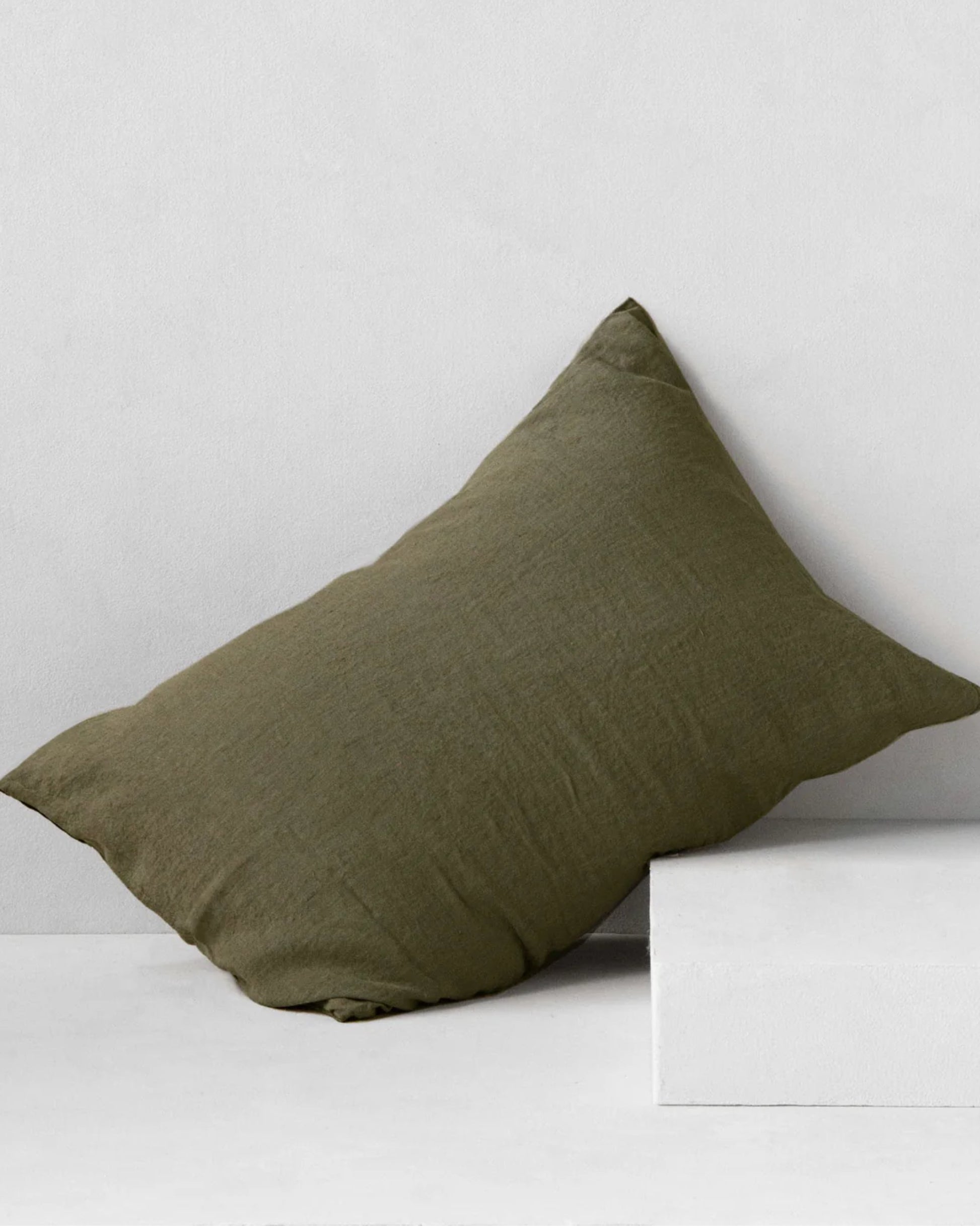 A BASIX STANDARD PILLOW CASE by Hale Mercantile Co in dark green is leaning against a light gray wall. It rests partially on a white rectangular block, enhancing the minimalist setting with neutral tones.