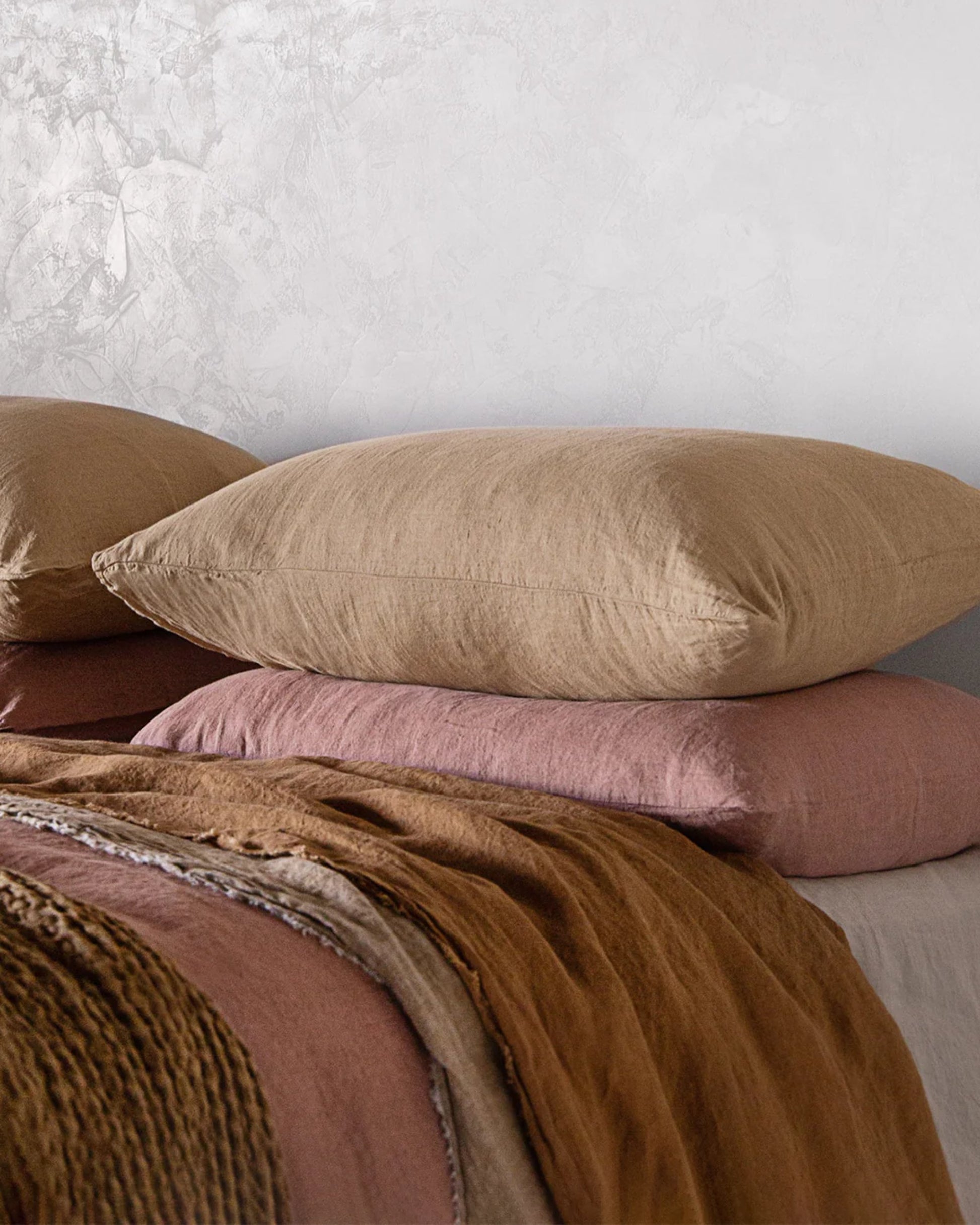 A stack of BASIX STANDARD PILLOW CASES by Hale Mercantile Co in brown and pink covers sits on a bed with matching linens. The wall in the background is textured and light gray, complementing the cozy, earthy tones of the bedding.