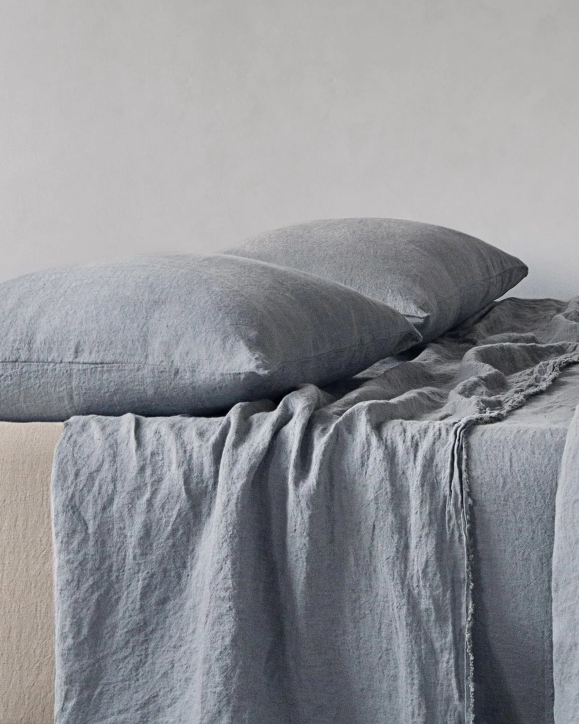 A neatly made bed features two large gray BASIX STANDARD PILLOW CASEs by Hale Mercantile Co, along with a matching gray linen blanket elegantly draped over a beige mattress. The bedding has a soft and textured appearance, enhancing the cozy and inviting ambiance against the plain, light-colored background.