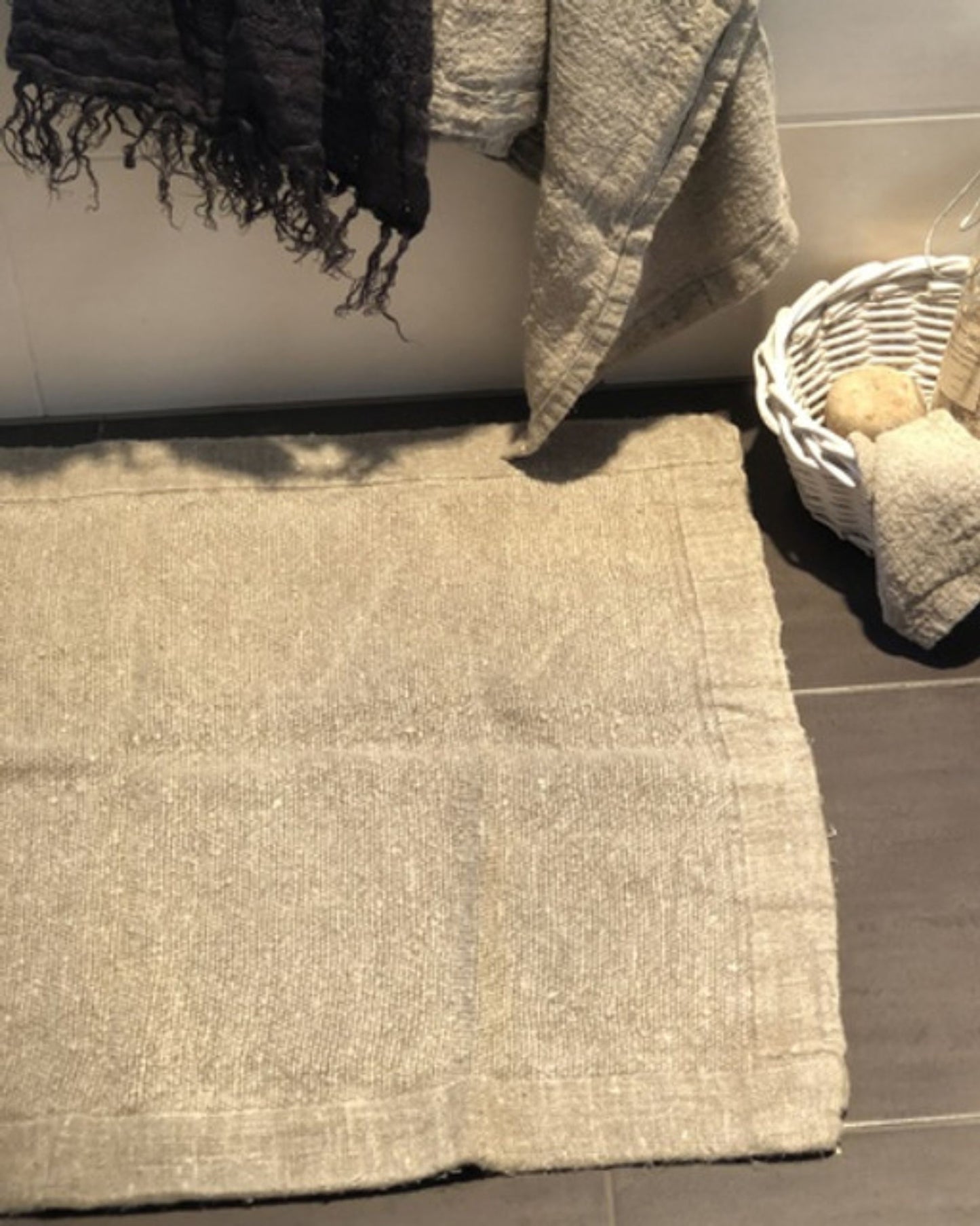 The dark tiled floor features the graceful presence of a BATH MAT - ALDER HANDLOOMED LINEN from Rustic Linen. Nearby, a white basket holds bath towels and a sponge, while textured towels hang with elegance over the edge of the tub, adding a luxurious touch to the bathroom decor.