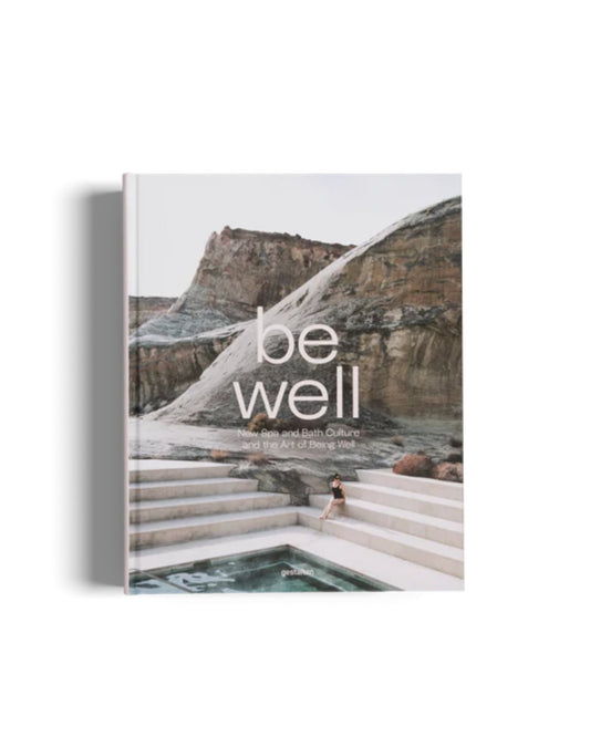 The image displays the cover of Flying Kiwi's "BE WELL - GESTALTEN," featuring a person sitting on steps near a pool in a rocky, natural landscape. Highlighting wellness, the cover text reads, "BE WELL: New Spa and Bath Culture and the Art of Being Well," providing insights into rejuvenating self-care practices.