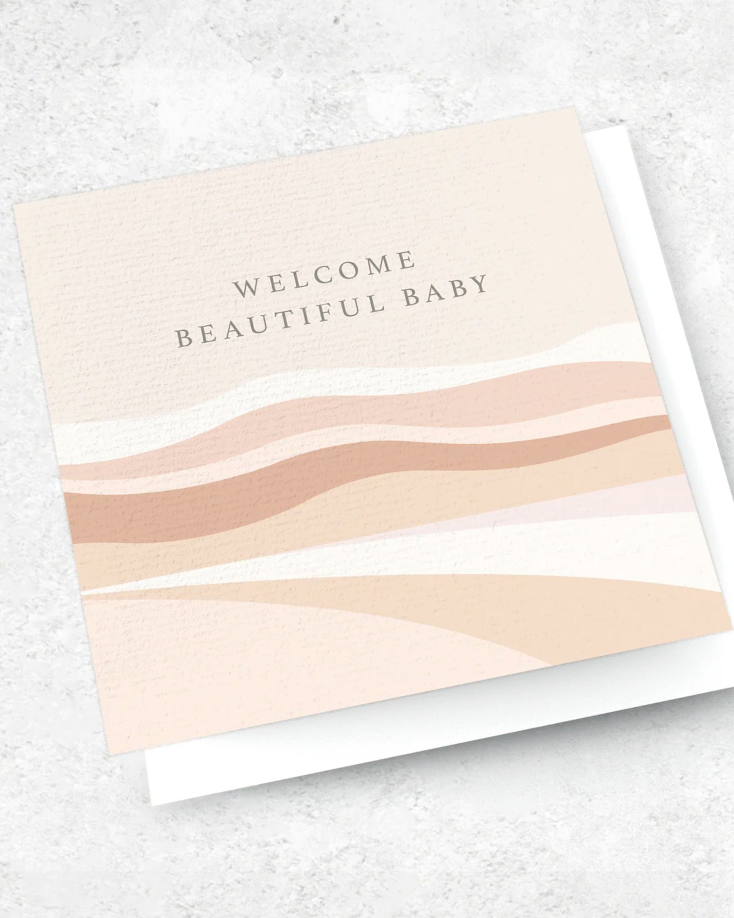 The "WELCOME BEAUTIFUL BABY - GREETING CARD" by Ink Bomb is a premium textured square card showcasing abstract beige and peach wave patterns. The phrase "Welcome Beautiful Baby" is elegantly centered at the top. It sits gracefully on a light gray marbled surface and comes with a 120gsm envelope.