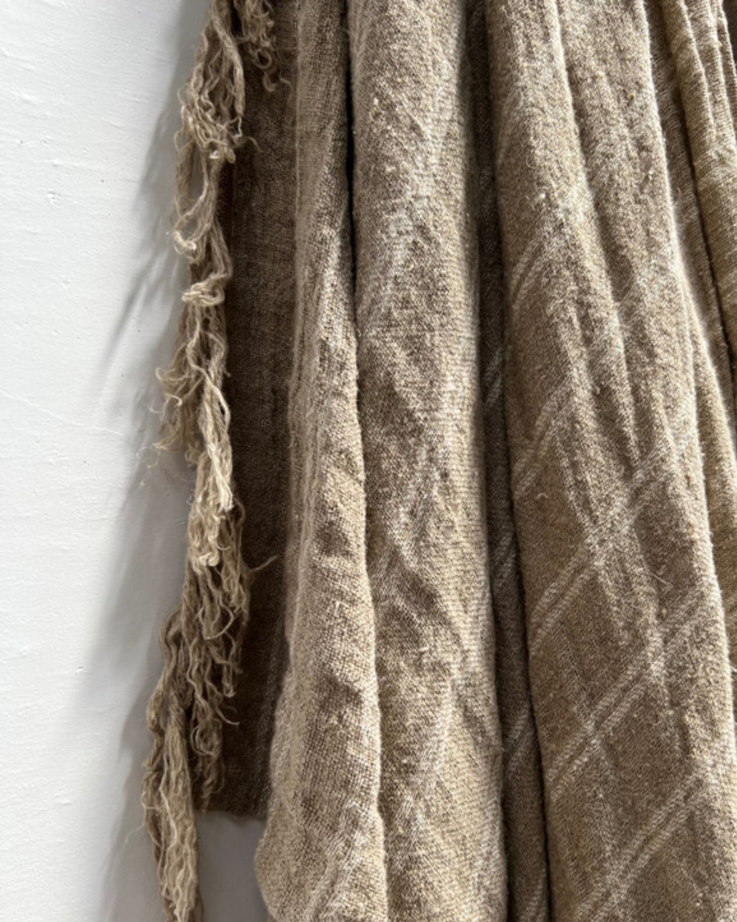 A close-up of a handloomed beige fabric, the Rustic Linen Bed Throw/Table Cloth with White Stripes, hangs against a white wall. Its plaid pattern and frayed edges exude rustic charm, enhancing any home decor with its cozy appeal.