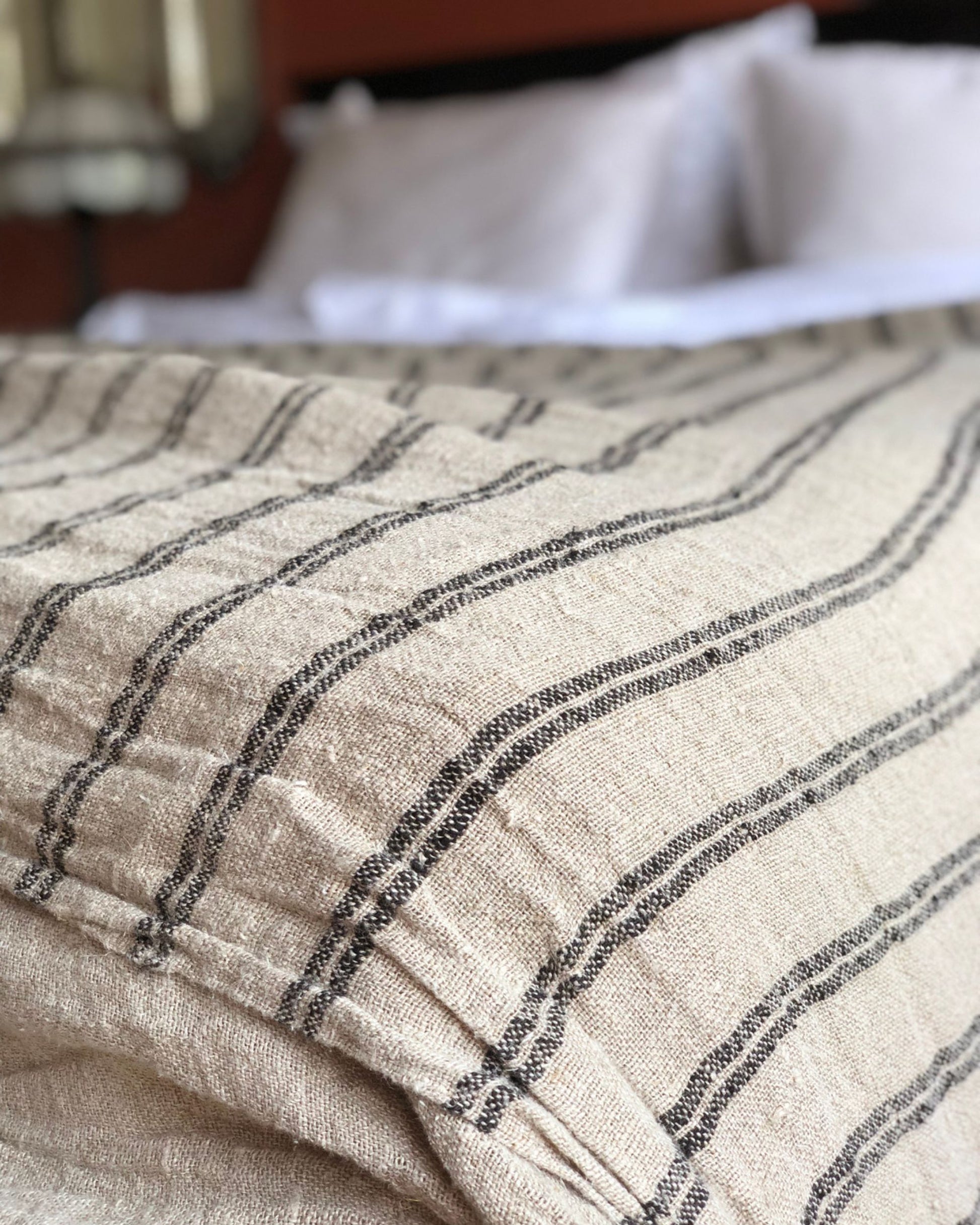 Close-up of a bed adorned with the BEDCOVER - BLACK STRIPE HANDLOOMED LINEN by Rustic Linen, showcasing its elegant black stripes. In the background, pillows and part of a headboard or wall can be seen, creating a cozy and inviting bedroom setting accentuated by the refined details of handloomed linen.