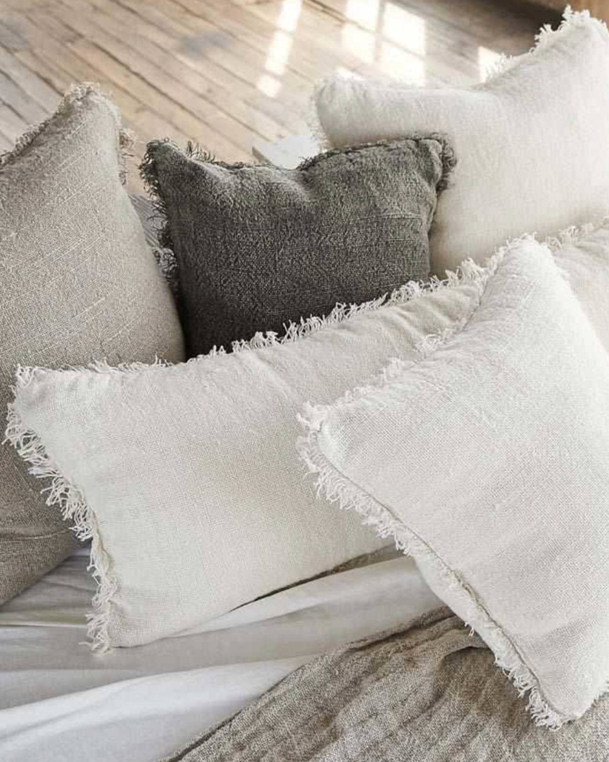 A cozy arrangement of textured pillows in neutral tones such as white, beige, and dark gray, featuring fringed edges. Among them is the Eadie Bedouin Linen Long Lumber cushion, adding a rustic chunky texture as they scatter on a light-colored bedspread in the sunlit room with a wooden floor.