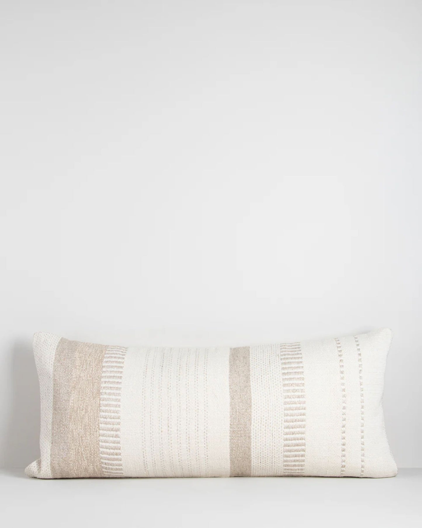 The BETTIE OATMEAL CUSHION by Baya is a lumbar-shaped throw pillow showcasing a minimalist design with horizontal stripes in diverse shades of beige and white on a simple background, epitomizing understated luxury.