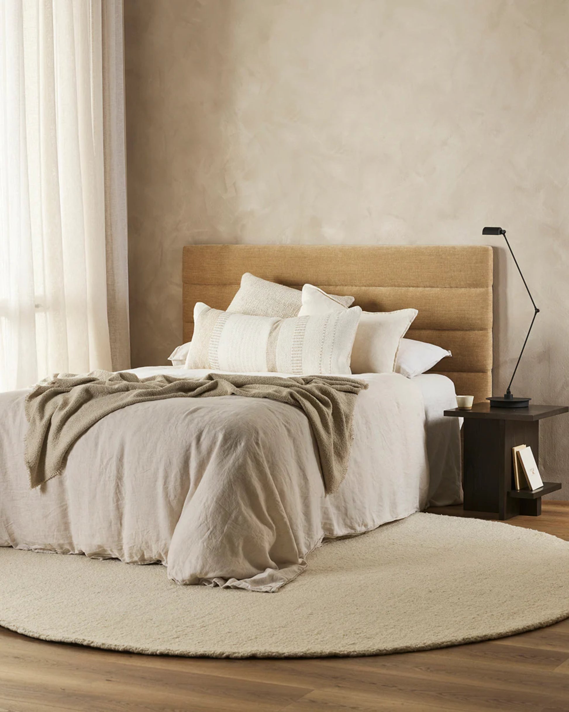 A cozy bedroom exudes understated luxury with its beige color scheme featuring the BETTIE OATMEAL CUSHION by Baya. The bed showcases pillows and an intricate weave blanket, complemented by a round beige rug. A wooden nightstand is adorned with a modern black lamp, while sheer curtains gently diffuse soft natural light throughout the room.