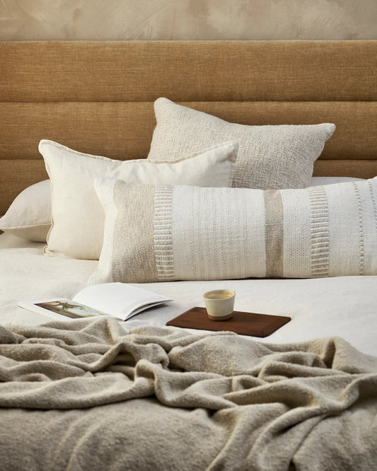 A cozy bed featuring Baya's BETTIE OATMEAL CUSHION and a beautifully woven blanket exudes understated luxury. An open book and a tray with a cup of coffee rest on the bed, creating an inviting atmosphere for relaxation.