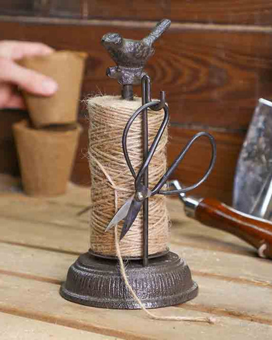 Topped with a charming rustic iron bird sculpture, the BIRD STRING HOLDER WITH SPIKE by CC Interiors artfully holds a spool of twine and scissors. Set against a wooden surface with softly blurred gardening tools and peat pots in the background, it's an ideal gift for any gardening enthusiast.