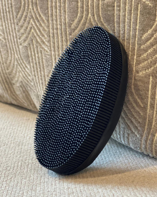 A close-up of the BLACK RUBBER TEXTILE BRUSH by Andree Jardin is shown resting against a beige textured cushion. The black, oval-shaped silicone brush features dense, short bristles, making it ideal for scrubbing or grooming and perfect for efficiently removing lint.