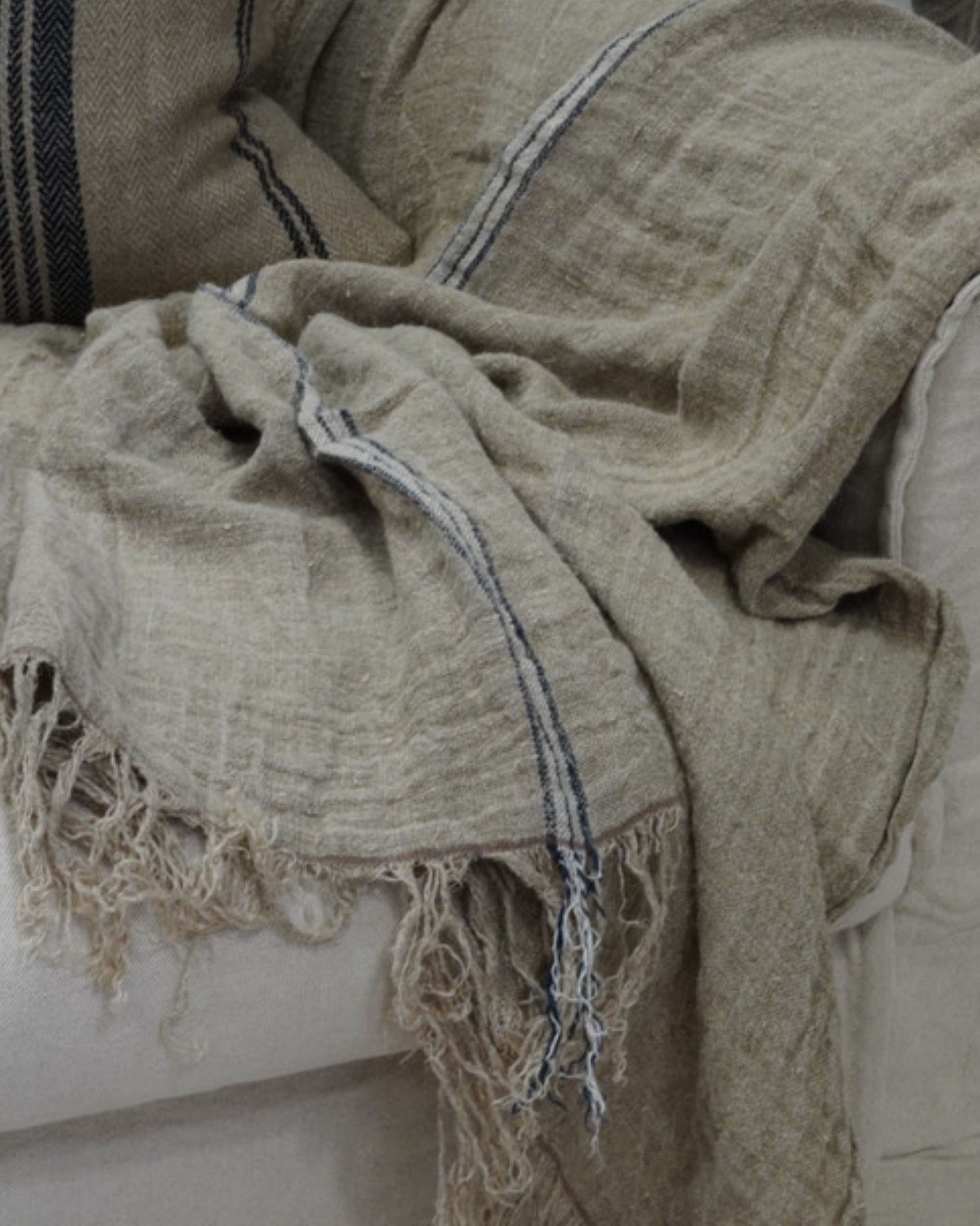 A cozy, textured Angaston Handloom Throw in a blue stripe design by Rustic Linen is draped over a plush, light-colored sofa. The edges of the handloom throw are frayed. A matching pillow with a blue stripe pattern is propped against the sofa.