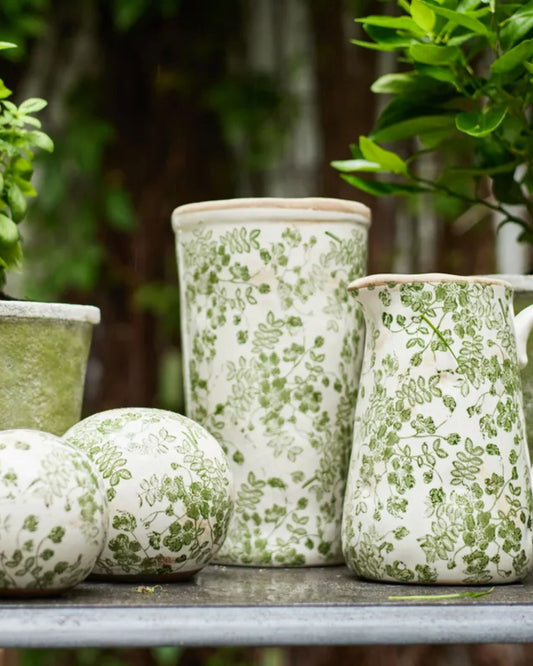 French Country's BOTANICAL VASE - GREEN FLORAL collection is elegantly arranged on a table, enveloped in lush greenery that crafts a tranquil garden scene brimming with rustic charm.