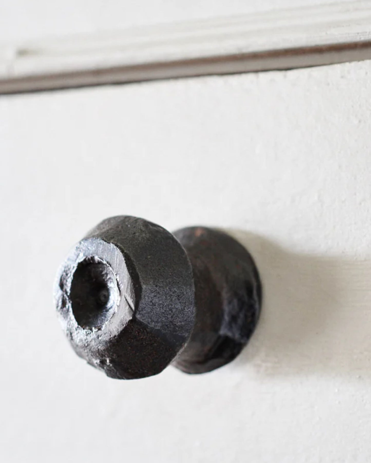 Close-up of a Society Ink Bower Wall Hook/Drawer Knob on a white door. The doorknob showcases a rustic and textured aesthetic, with its dark finish reflecting artisanal craftsmanship and visible signs of wear.