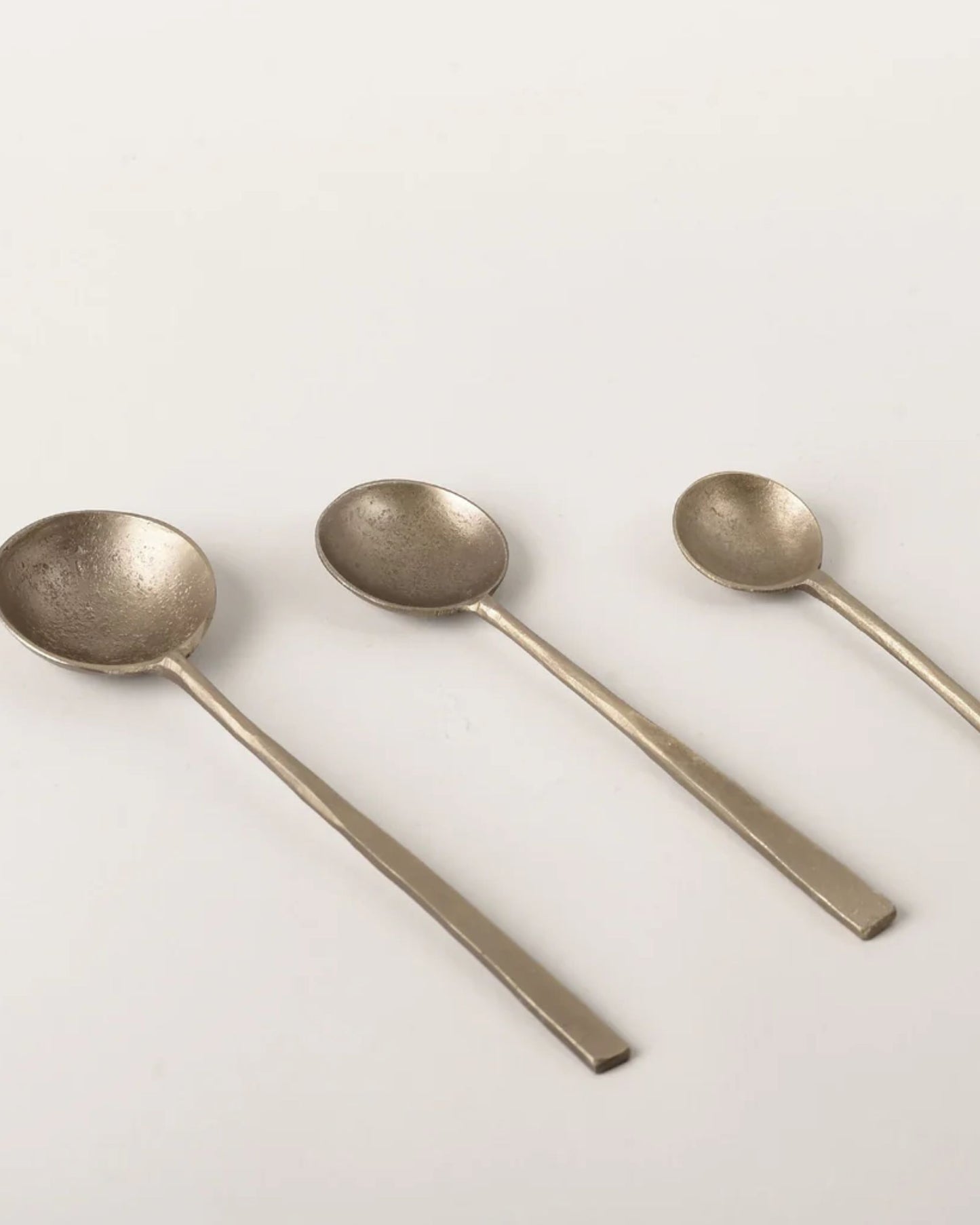 A trio of spoons from the New Direction brand, crafted in the ANTIQUE BRASS SPOON style, are displayed in a row. These metallic pieces feature a vintage-inspired design with a brass finish, each showcasing a round bowl and straight handle against the light surface. Arranged from largest to smallest, they exude an appealing rustic charm.