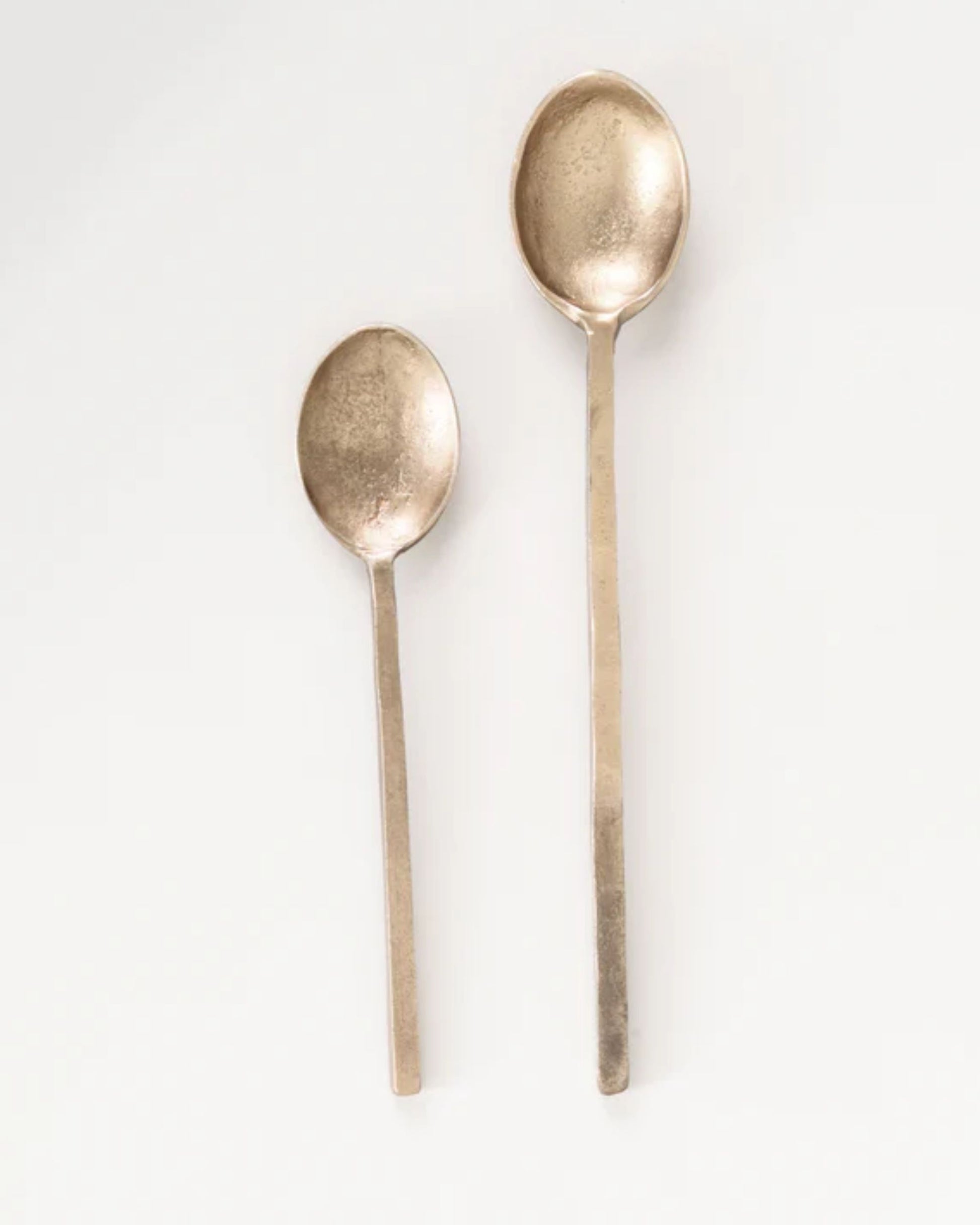 Featuring a larger and a smaller spoon on a white background, the BRASS SPOON WITH SQUARE HANDLE by New Direction exhibits square handles and a slightly rustic finish. These spoons offer minimalist and modern elegance, enhancing any dining experience.