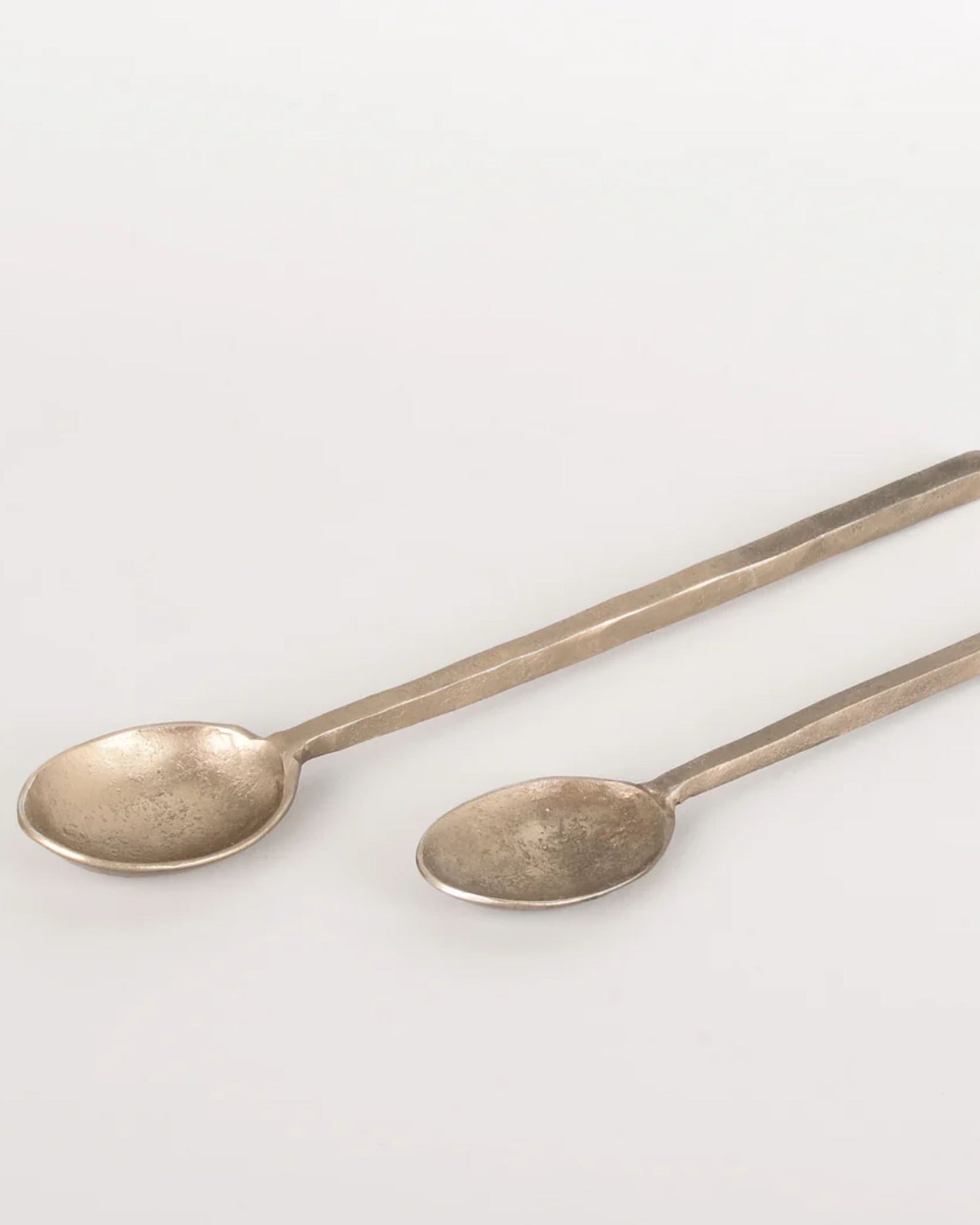 On a light gray surface, two BRASS SPOON WITH SQUARE HANDLE pieces from New Direction, each varying in size and featuring a rustic bronze tone, rest side by side. Their handcrafted appearance and subtly textured square handles exude modern elegance.