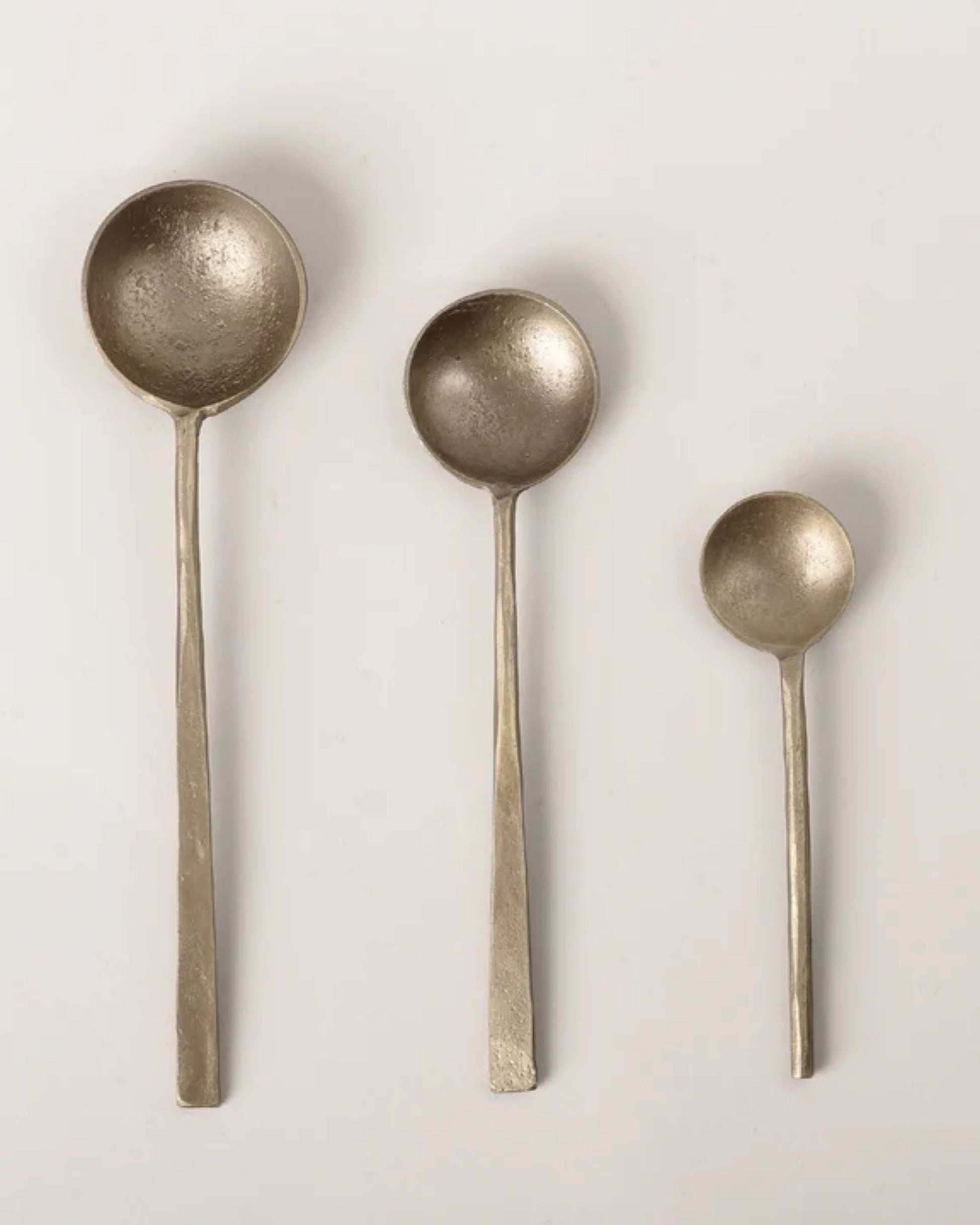 Three ANTIQUE BRASS SPOONS by New direction, each of a different size, are arranged vertically against a light background. These spoons, echoing a vintage-inspired design, showcase a rustic, textured brass finish and feature long, slender handles.