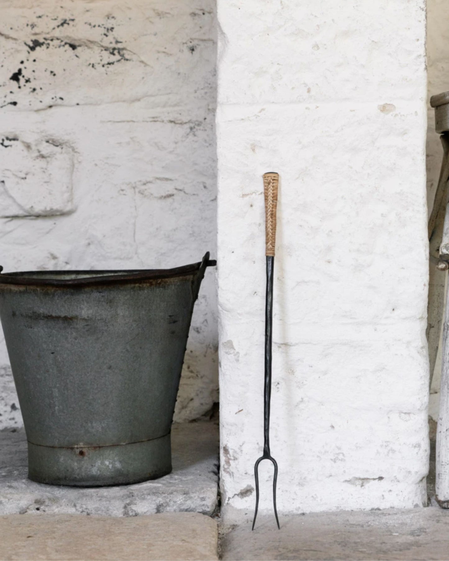 A rustic scene unfolds with Society Ink's CAMPFIRE FORK, a long-handled tool featuring a woven grip and paired with a metal bucket against a whitewashed brick wall. This glamping fire tool evokes a cozy, farm-like atmosphere, making it perfect for enhancing outdoor adventures or serving as the ideal camping gift.