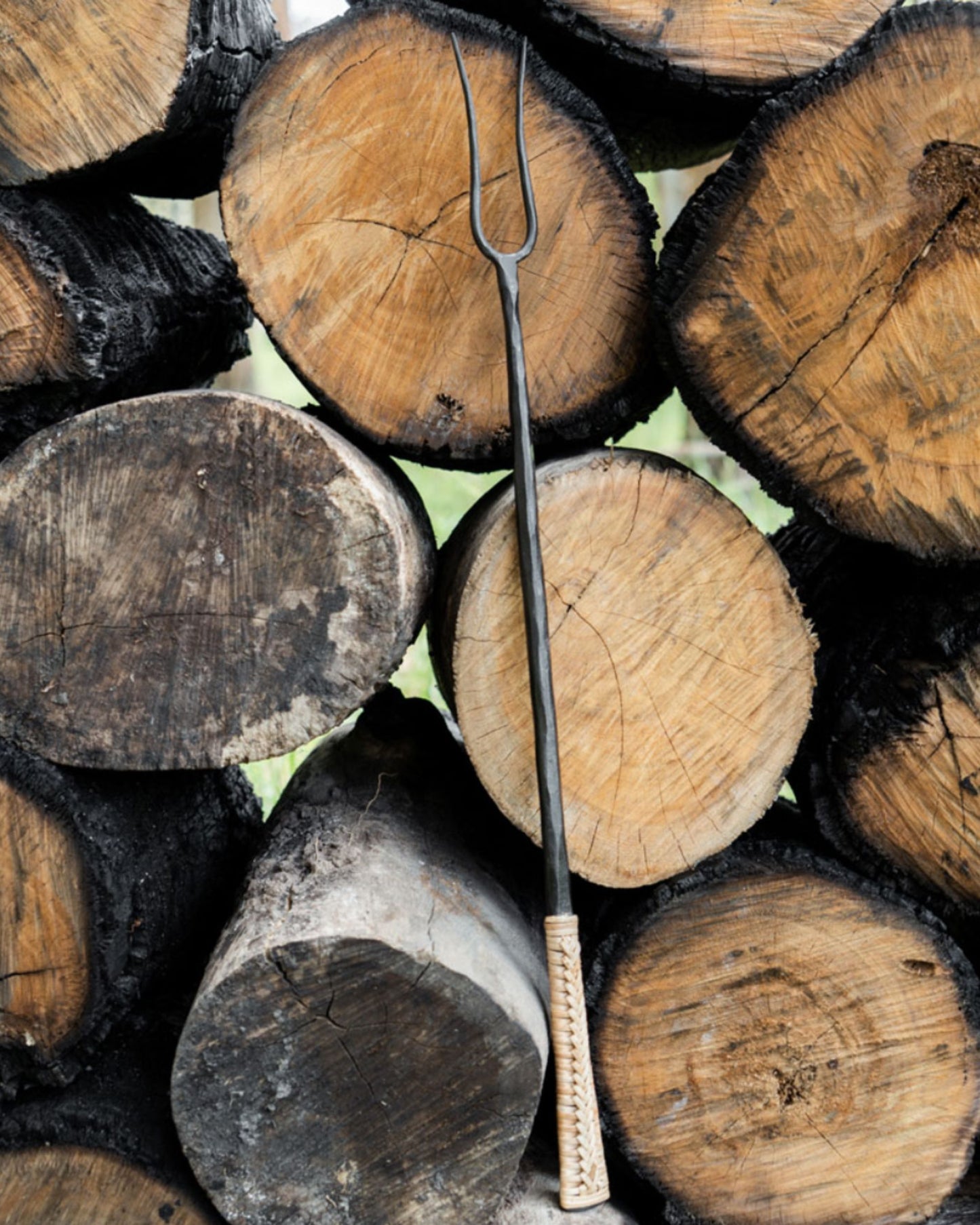 A Society Ink CAMPFIRE FORK with a wooden handle leans against a stack of neatly arranged firewood logs, offering the perfect glamping fire tool. The rough, circular cut ends of the logs display distinct growth rings and textures, creating a rustic ambiance ideal for the discerning glamper.