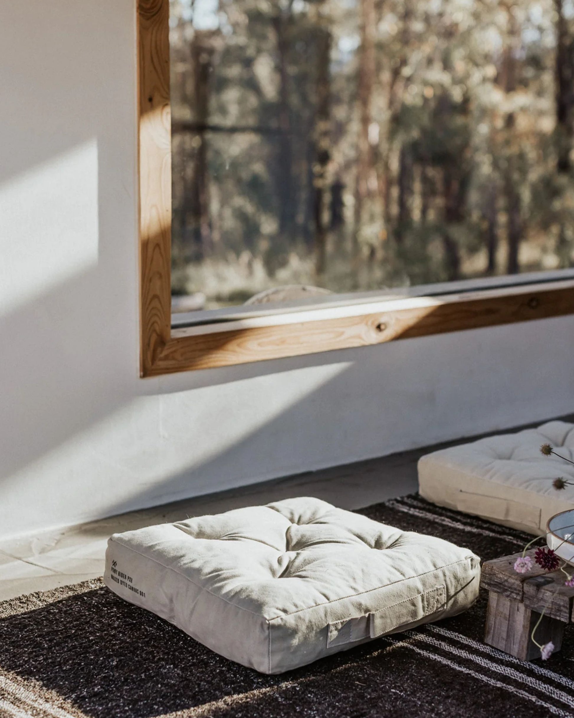 A cozy room features the CAMP FIRE OUTDOOR FLOOR CUSHION - ANGORA by Pony Rider, resting on a woven rug in front of a large wooden-framed window. Sunlight floods in, accentuating the earthy tones of the decor and providing a view of trees outside.