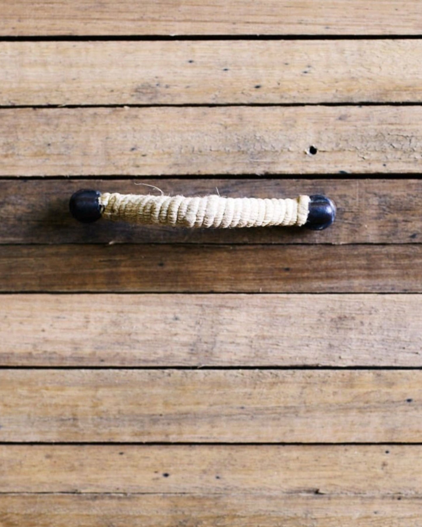 Introducing the CAPTAINS DRAWER PULL by Society Ink—a rustic wooden handle wrapped in rope, evoking seafaring themes. Mounted on a wooden surface with horizontal planks, it features dark end caps that enhance its simple, natural aesthetic. This distinctive piece is perfect for home decor enthusiasts who seek unique touches such as rope drawer pulls.