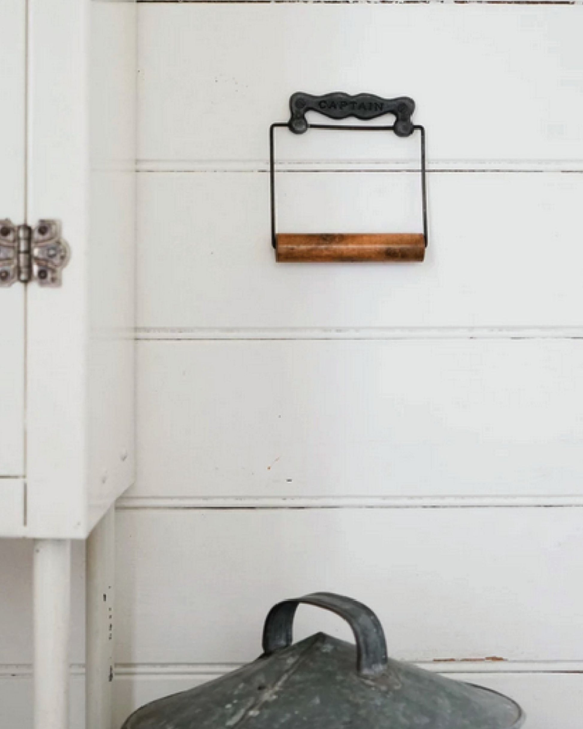 Vintage kitchen scene featuring a white wooden cabinet on the left and a rustic metal and wood paper towel holder from Society Ink, known as CAPTAINS ROLL, mounted on a white wall. A galvanized metal lid is placed below, enhancing the farmhouse charm of this delightful home decor setup.