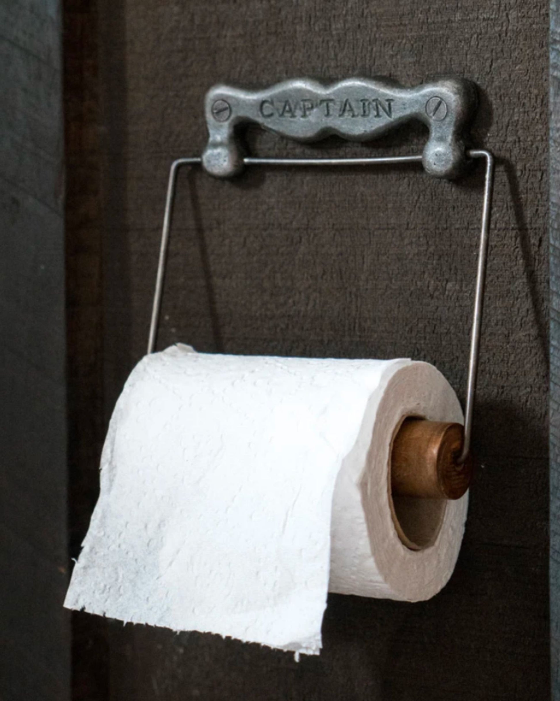 A roll of toilet paper hangs from a vintage metal holder with "CAPTAINS ROLL" by Society Ink elegantly engraved on it, adding a touch of pirate flair to the rustic home decor. The holder is fastened to a dark wooden wall, and the textured paper roll is partially unraveled.