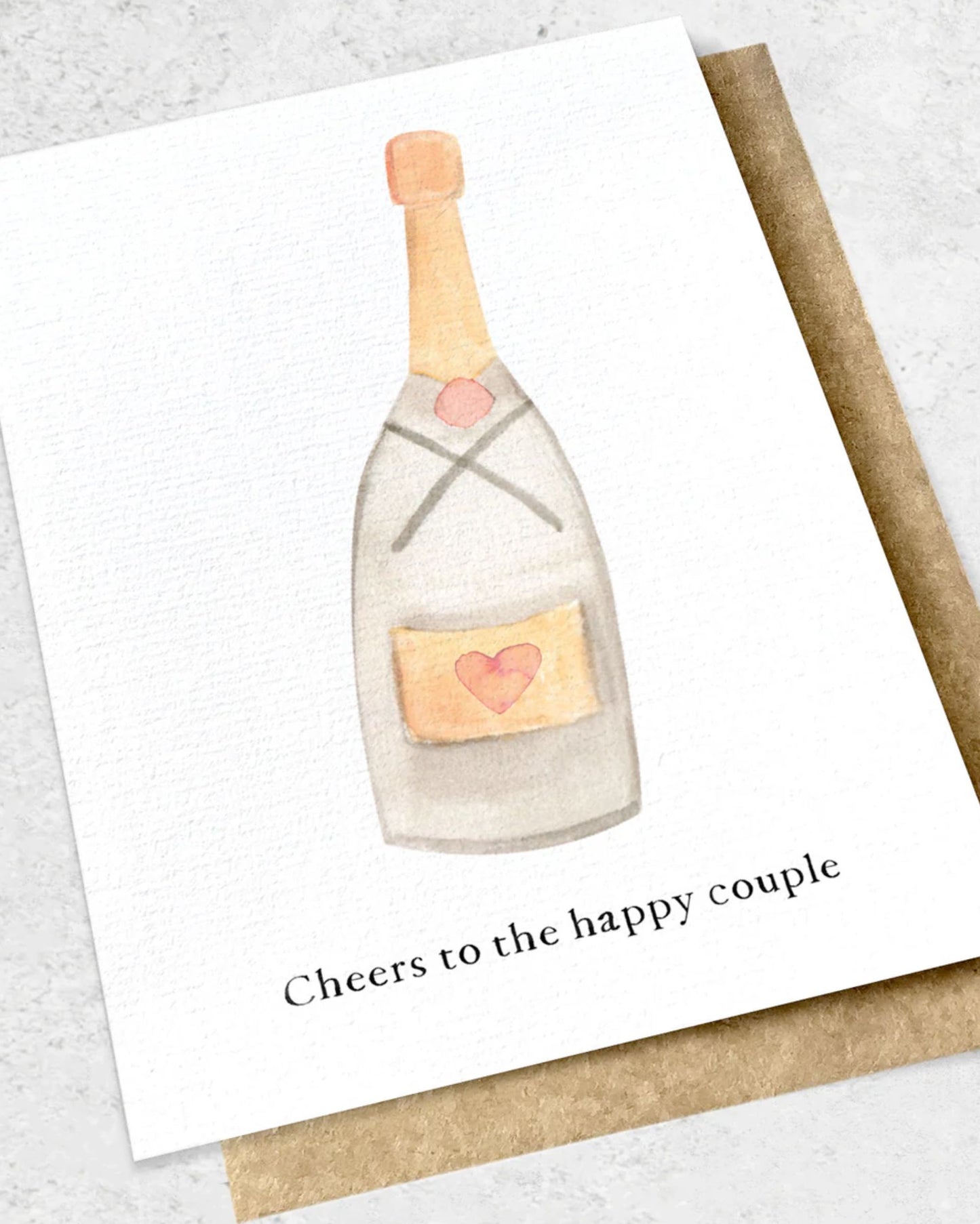 An Ink Bomb premium textured greeting card, the CHEERS TO THE HAPPY COUPLE design features a watercolor illustration of a champagne bottle adorned with a pink heart and tan label. The phrase "Cheers to the happy couple" is artfully inscribed below. This card is presented on a kraft envelope, elegantly resting on a light surface.