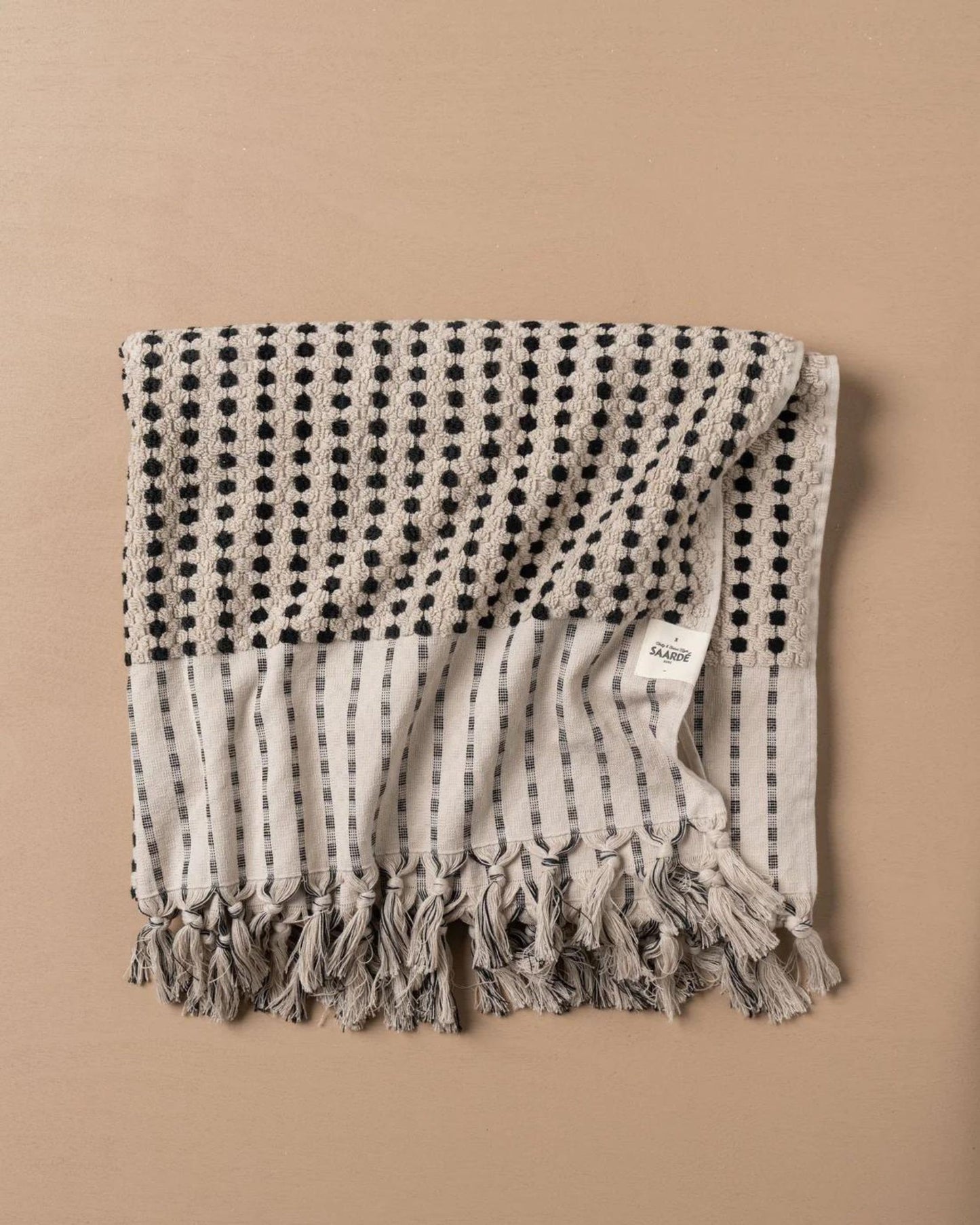 A CHICKPEA BATH SHEET from Saarde, featuring a beige and black patterned design, is spread out on a beige surface. Made from Turkish cotton, it showcases polka dot and striped patterns with a fringe, and a white tag displaying text can be seen.