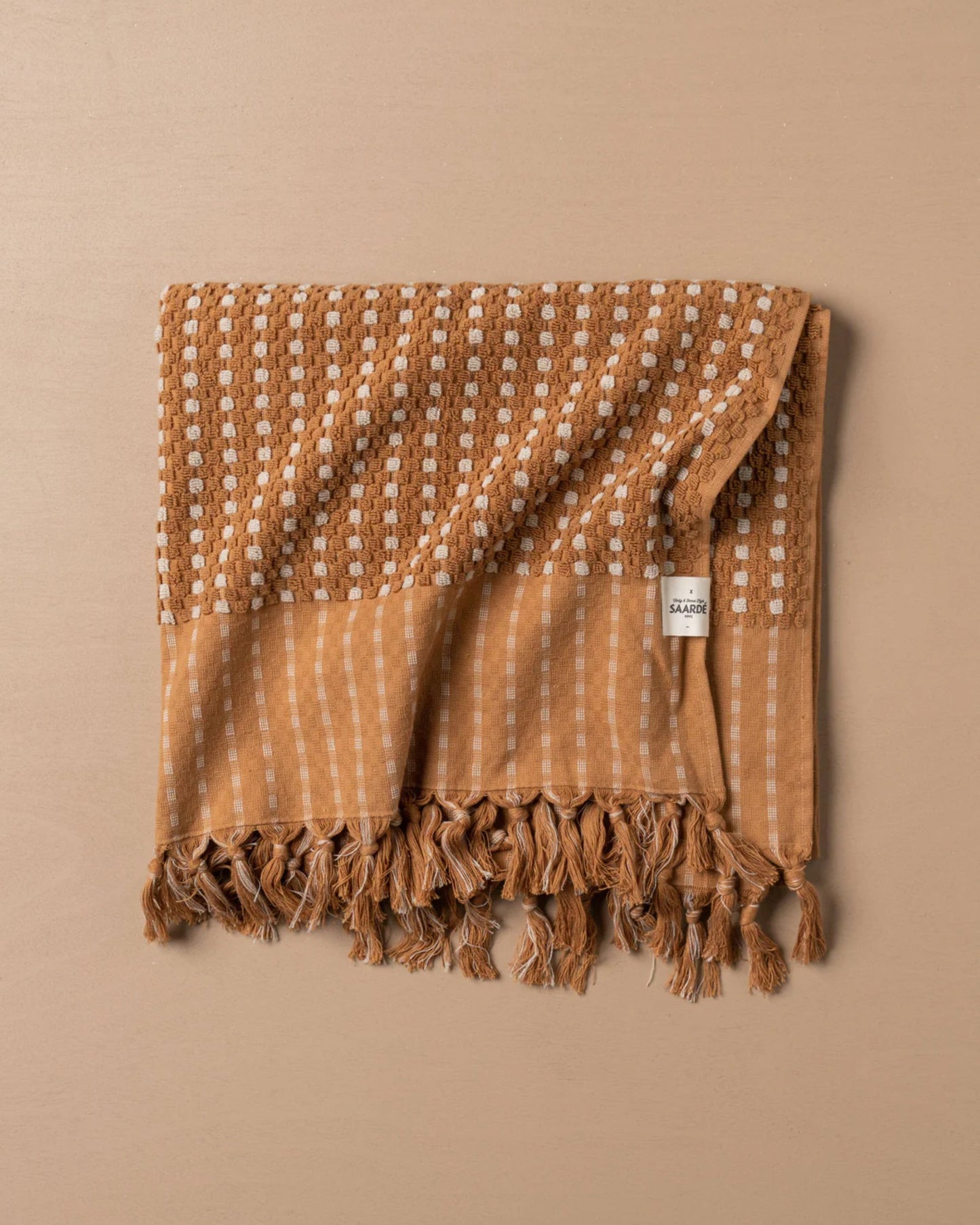 A CHICKPEA BATH SHEET by Saarde, crafted from super absorbent Turkish cotton, features a textured surface in brown and beige with a pattern of squares and lines. The fringed edge adds elegance to the design, complemented by a small white tag.